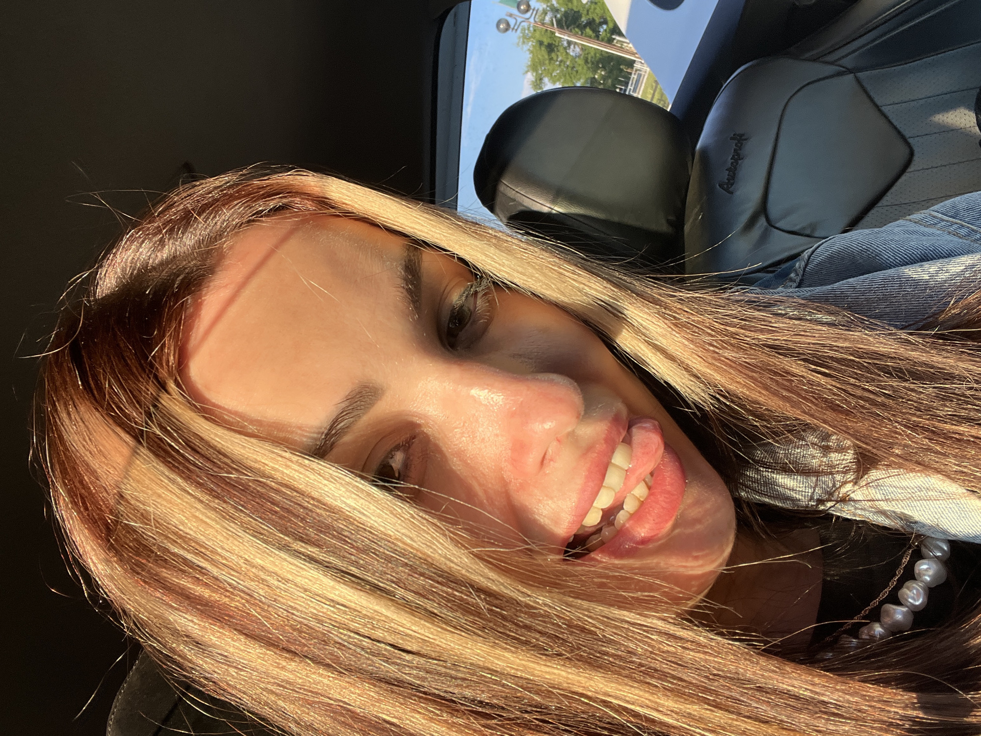 A woman is in a car in Unknown, Unknown. The woman has long hair with blond highlights. She is wearing a denim jacket and a pearl necklace. The car seat is black leather.