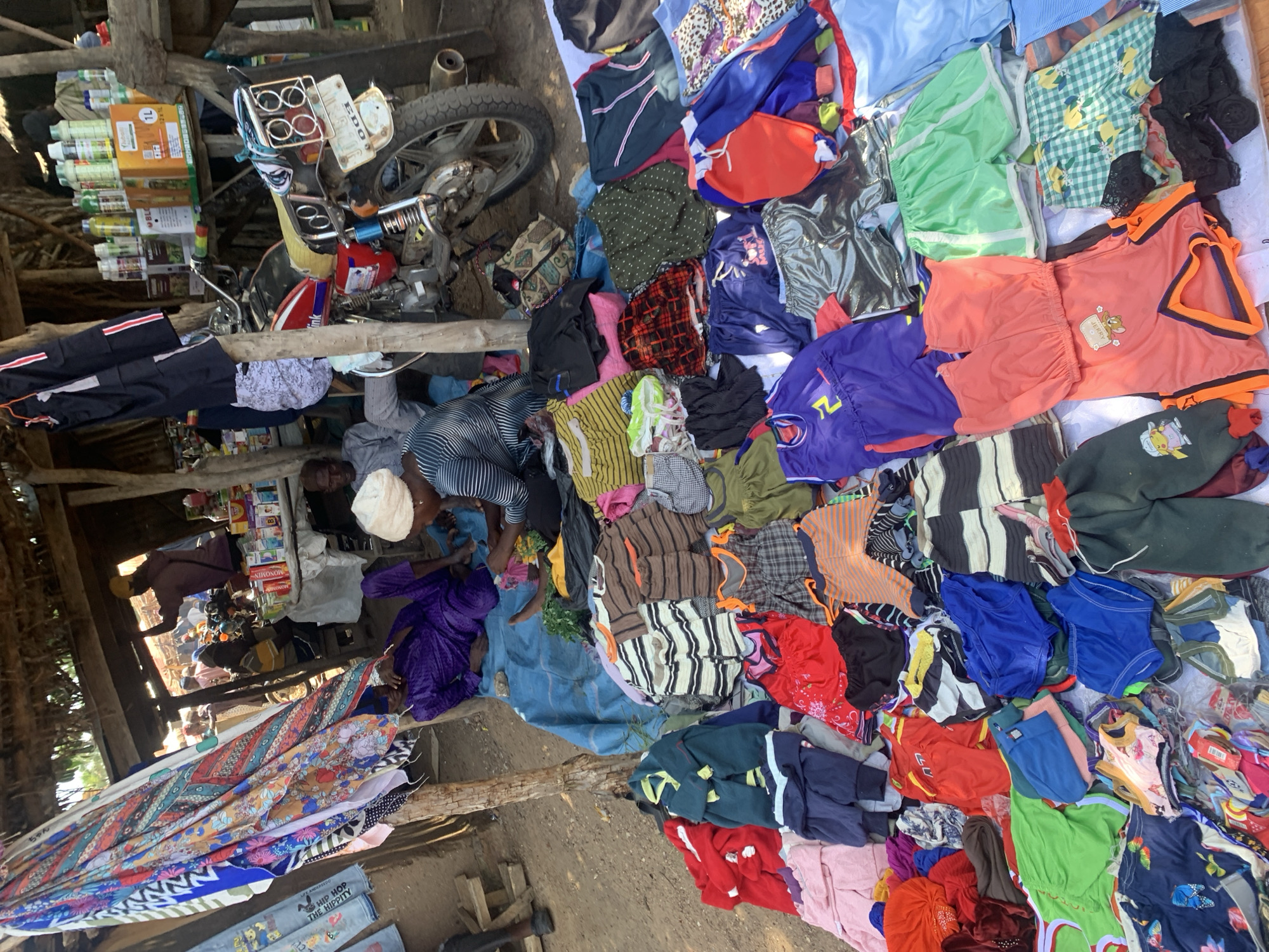This is a medium-shot, high-angle photograph taken outdoors, likely in Boi A, Nigeria. The scene depicts a bustling open-air market stall overflowing with a colorful assortment of clothing. In the foreground, a large pile of folded fabrics, featuring vibrant geometric patterns and floral motifs, is displayed prominently.
Dominating the mid-ground is a motorcycle, its rear wheel and part of its frame visible, positioned amongst stacks of clothing. To the left, shelves are laden with numerous bottles, some appearing to be pesticides or agricultural products, with prominent text including "1L" and other illegible labels.
Several individuals are present in the scene. In the foreground, a person wearing a striped shirt is partially visible. In the mid-ground, a person in a purple garment is seen. Further back, a person in a white cap and another in a dark shirt are interacting near the shelves of bottles. The overall impression is that of a busy marketplace, possibly in the morning or afternoon given the natural lighting. The ground appears to be dirt. The arrangement of goods suggests a commercial activity of selling apparel and other wares.