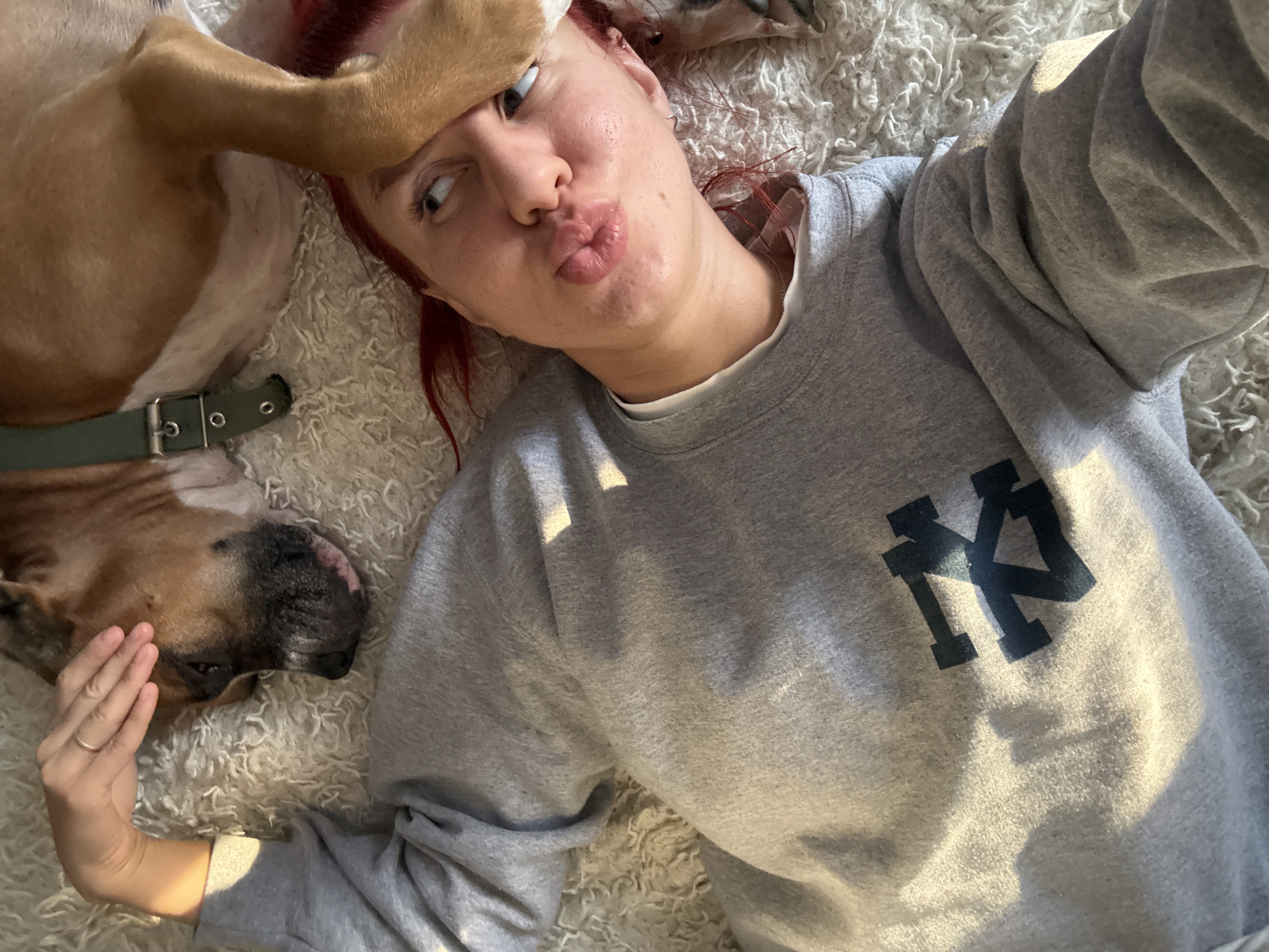 A person with red hair is lying on a fluffy white rug, making a kissing face at the camera. A dog is lying next to the person, with its paw resting on the person's face. The person is wearing a gray sweatshirt with a dark blue logo. The dog is brown and appears to be a pit bull or similar breed, wearing a green collar. The scene appears to be indoors, possibly a living room. There is no information to suggest this is in Rostov-on-Don, Russia.
