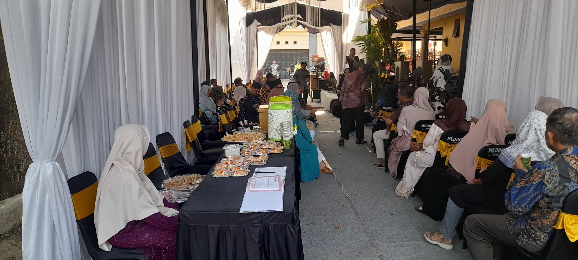 The image depicts an outdoor event in Taman, Indonesia, characterized by a row of white drapes on the left and a covered area on the right with yellowish awnings. A group of people are seated on chairs arranged in rows, facing a central area where a person appears to be speaking into a microphone. In the foreground, a table covered with a black tablecloth is laden with food items, including small pastries and snacks in clear plastic containers. A green and white patterned dispenser, along with stacks of paper cups, are also visible on the table. Several individuals are wearing headscarves, suggesting a cultural or religious context. The ground appears to be paved. The overall scene suggests a gathering, possibly a reception or a public event.
