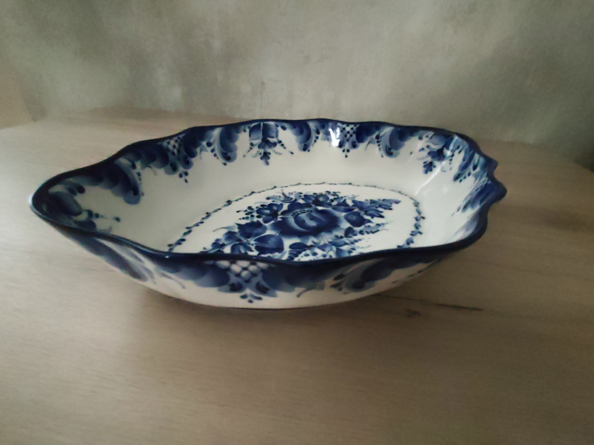 A white oval-shaped ceramic bowl with a scalloped rim is positioned on a light-colored wooden surface. The bowl is decorated with a blue floral pattern. The background is a light gray, textured surface. There are no people or animals present. The image does not contain any recognizable landmarks or features that would indicate a specific location in Sochi, Russia.