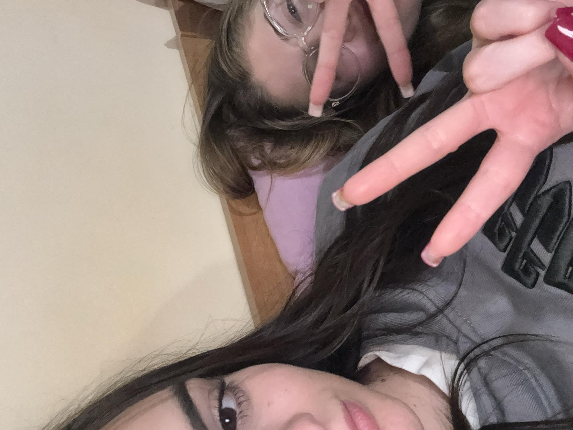 Two young women are posing for a selfie. One woman has long, dark hair and is wearing a grey jacket over a white shirt. The other woman has shoulder-length brown hair and is wearing round glasses. Both women are making the peace sign with their hands. The background is a plain wall. The location is likely indoors.