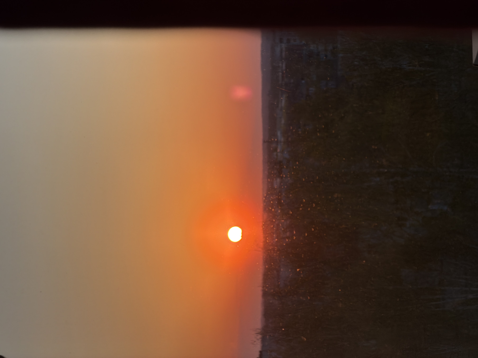 The image shows a sunset viewed through a window in Chaikovsky, Russia. The left side of the image depicts a hazy, orange sky transitioning into a bright orange sun near the center. The right side reveals a dark, partially visible urban landscape, likely a residential area, obscured by the window pane. Droplets are visible on the window. The top portion of the image features a blurred dark frame, possibly a window frame.