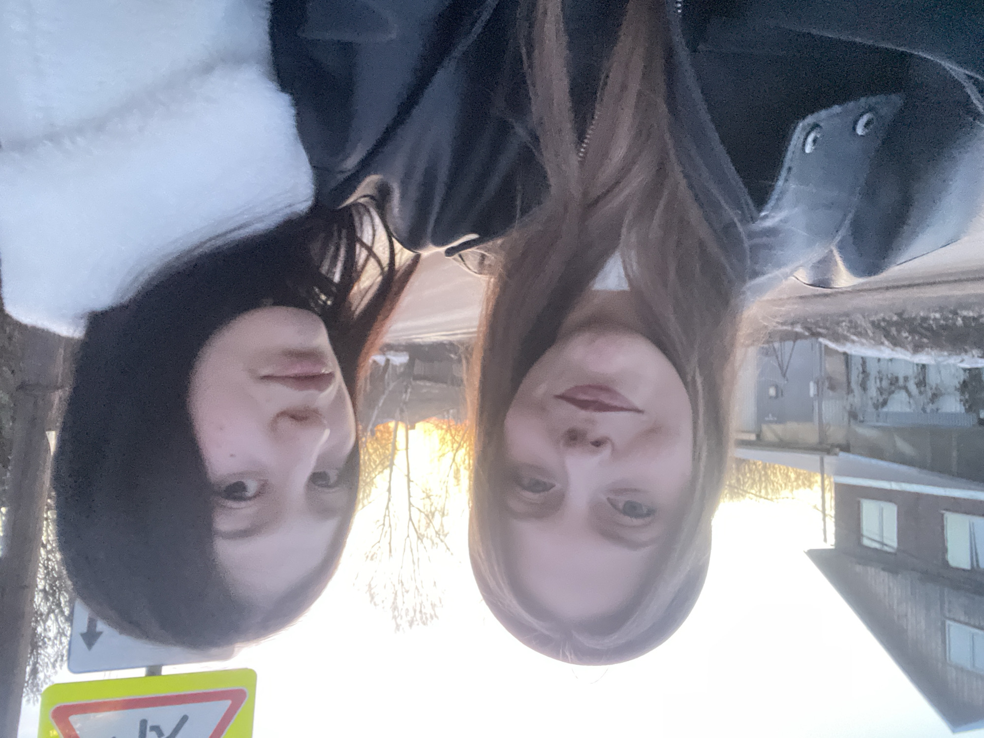 Two young women are posing for a selfie outdoors in Фоки, Russia. The woman on the left has dark hair and is wearing a jacket with a white fur collar. The woman on the right has light brown hair and is wearing a black leather jacket. They are standing on a street with a road sign to their left. In the background, there are trees, houses, and the setting sun.