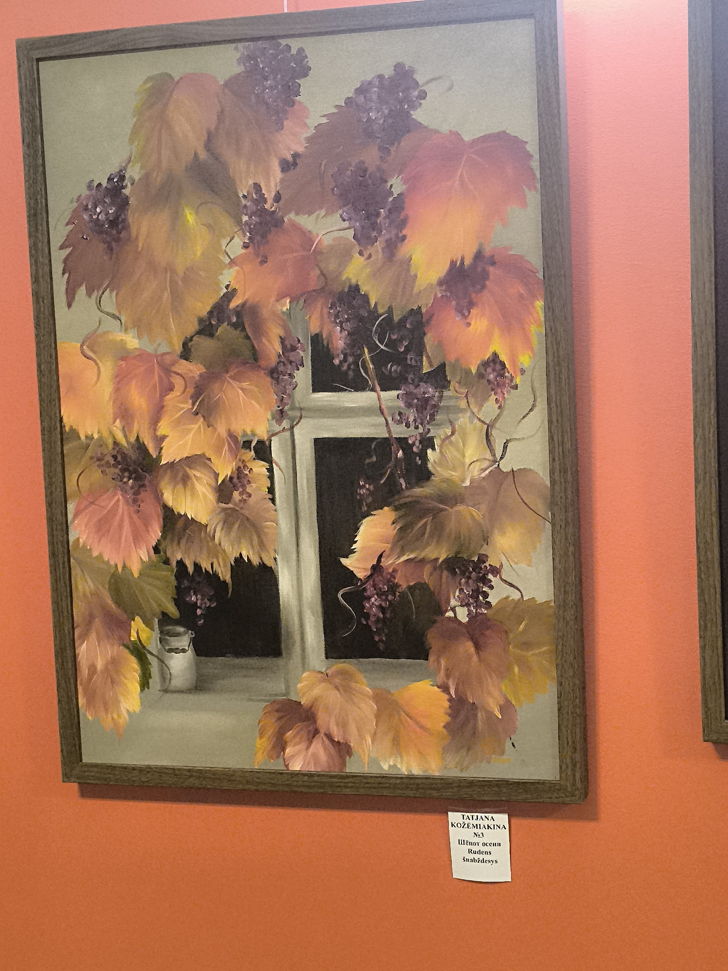 The image shows a painting of grapevines with purple grapes and yellow and orange leaves, partially obscuring a window. The painting is framed in a dark wooden frame and is displayed on an orange wall. A small white label is attached to the wall below the painting, containing text that appears to be a title and artist's name in Cyrillic and Latin script, along with a word that may be in Latvian. The text on the label reads "TATJANA KOZEMIAKINA N°3 Шёлк осени Rudens Šmālždesys". This suggests the artwork is likely on display in Klaipėda, Lithuania, as this is a port city where various art exhibitions are held. The overall scene is that of an art exhibition or gallery. There are no people or specific actions depicted in the image itself.
