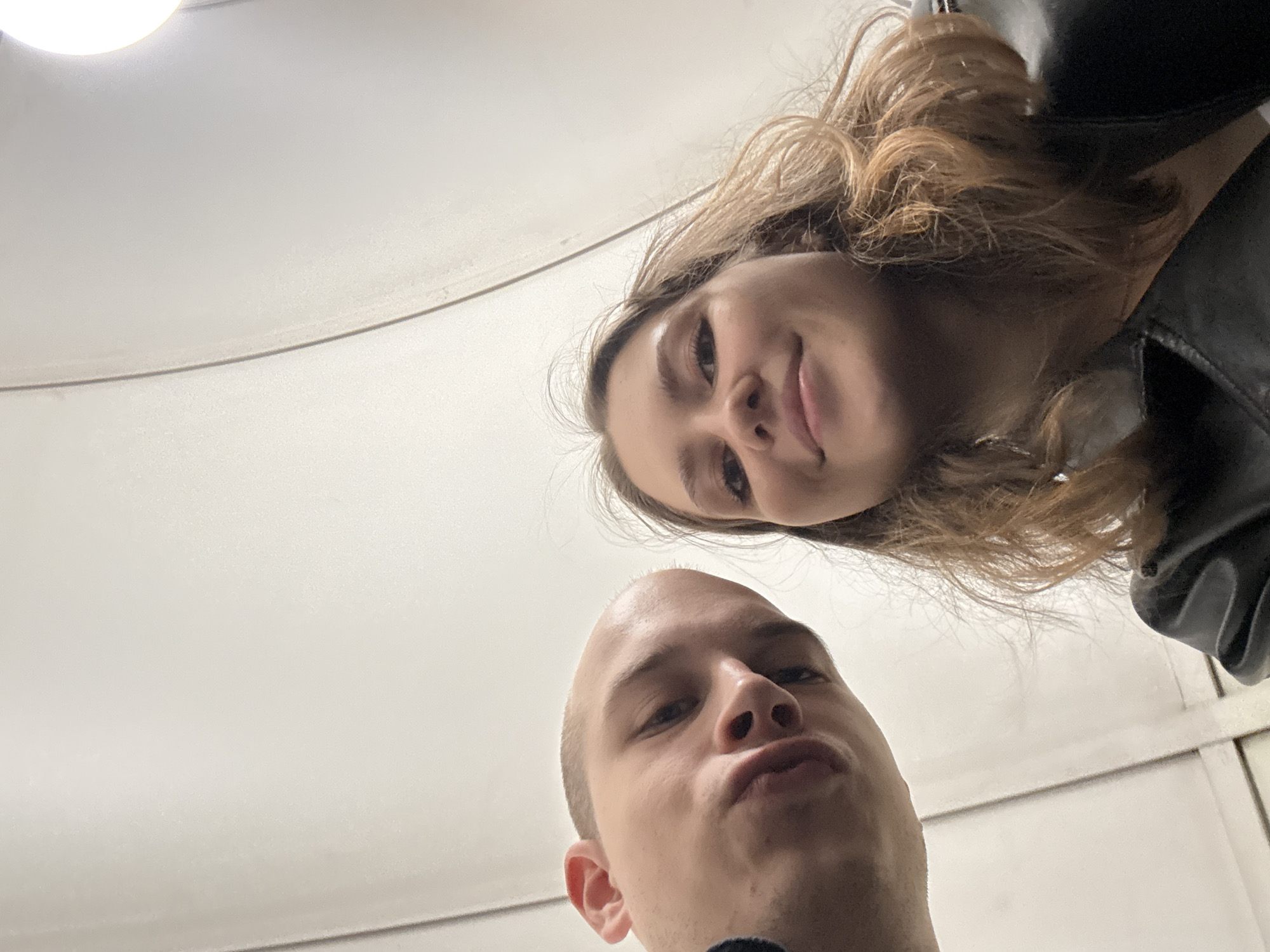 This is a low-angle, close-up selfie of two individuals, a man and a woman, appearing to be in an urban, possibly public, indoor location. The background is a smooth, curved, white ceiling with a visible seam or joint running vertically. To the right, a bright light source is partially visible.
The man, on the left, is seen from the forehead down, with his head tilted downwards. He has a shaved head and his face is partially obscured by shadow. His lips are pursed. He is wearing dark clothing, possibly a sweater or jacket, on his left shoulder and neck.
The woman is on the right, also looking upwards at the camera. She has shoulder-length, wavy, light brown hair. She is smiling and her eyes are open. She is wearing a dark, possibly leather, jacket over a lighter colored garment.
The perspective suggests the photo was taken from a low vantage point, looking up at the subjects. The overall lighting is bright and diffused, with a warm tone. The clean, seamless background and artificial lighting suggest an indoor environment such as a metro station, an airport terminal, or a modern building interior in Saint Petersburg, Russia.