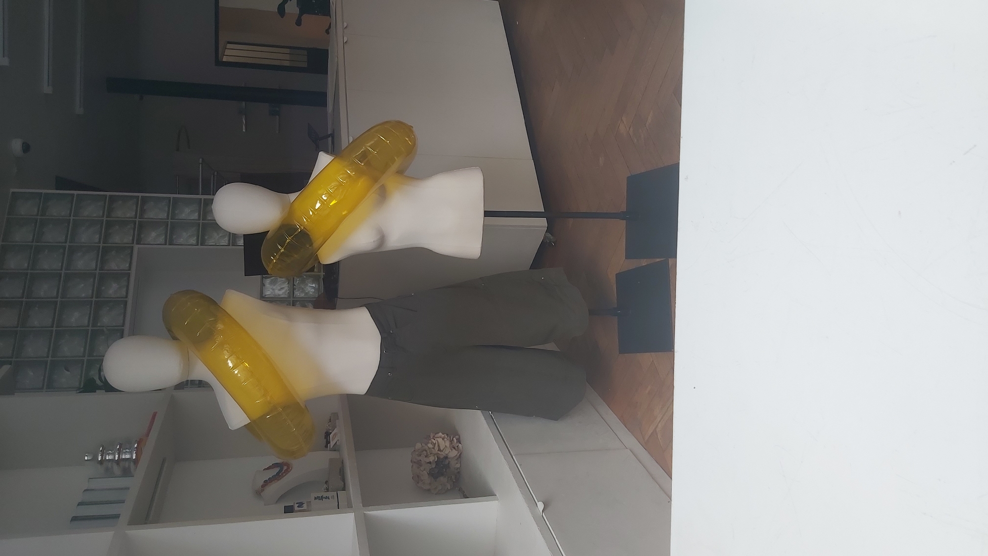 This is a photograph taken from inside a store, looking out through a window. Two mannequins are displayed in the foreground, both wearing large, yellow, inflatable objects around their torsos. The mannequin on the left is wearing dark green, wide-legged capri pants and has its arms behind its back. The mannequin on the right is a bust form and is also wearing the yellow inflatable object. The store interior features white shelving units with various items displayed, including jewelry on a stand and some decorative items. In the background, there is a wall with frosted glass tiles, a counter area, and a door. The lighting suggests it is daytime. The photograph appears to have been taken in Paris, France, indicated by the presence of a camera security device on a ceiling fixture.