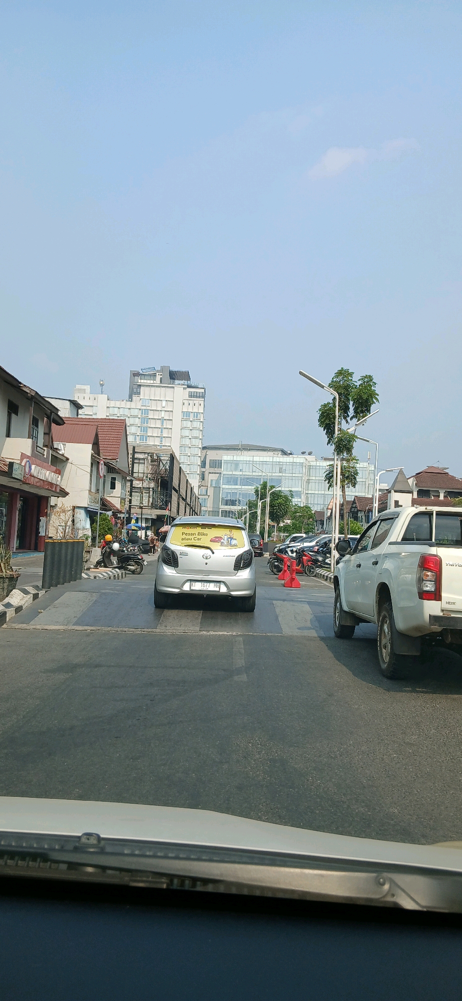 The image captures a street scene in Samarinda, Indonesia, likely during daylight hours with clear skies. In the foreground, a silver car is stopped at a pedestrian crossing. The car has a sticker on its rear window that reads "maxim Pesan Bike atau Car". To the right of the silver car, a white pickup truck is also stopped. Behind the silver car, several motorcycles are visible, some with riders. The street is paved, and there are buildings on both sides of the road. On the left, there are low-rise commercial buildings with awnings. On the right and in the background, larger modern buildings, including a multi-story hotel or office building with a glass facade, dominate the skyline. Streetlights and trees are also present along the road. The overall impression is of a bustling urban environment.