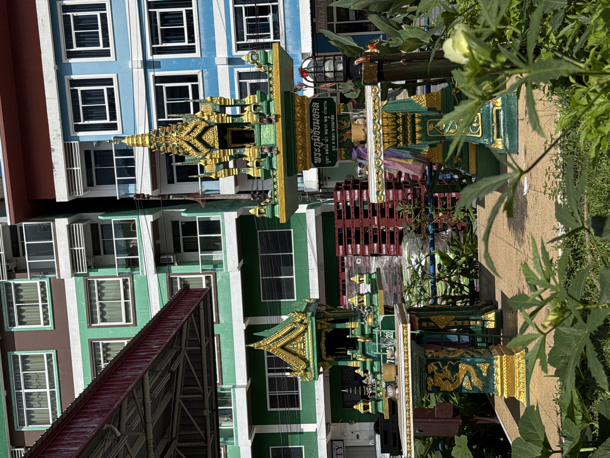 In the city of Patong, Thailand, an outdoor scene features two ornate spirit houses, or San Phra Phum, placed on a tiled patio. The spirit houses, predominantly green with gold accents, are adorned with intricate carvings and layered roofs. The larger spirit house, on the right, has a sign in Thai script on its base. Both structures are surrounded by lush green foliage. In the background, a multi-story building displays a colorful facade of brown, green, and blue sections, punctuated by numerous windows. Red and white pallets are stacked to the left of the larger spirit house. The lighting suggests it is daytime, with sunlight casting shadows.