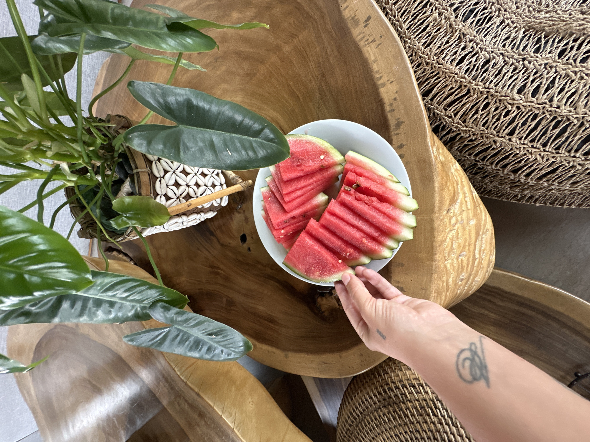 From an elevated perspective, the scene depicts a wooden coffee table in Sukawati, Indonesia, adorned with several objects. A small plant, situated in a basket decorated with seashells, sits on the left side of the table. To the right, a white bowl contains several slices of watermelon. A person's hand reaches for a slice, their arm visible with a tattoo on the inner forearm. Additional woven furniture pieces are partially visible along the right edge of the frame.