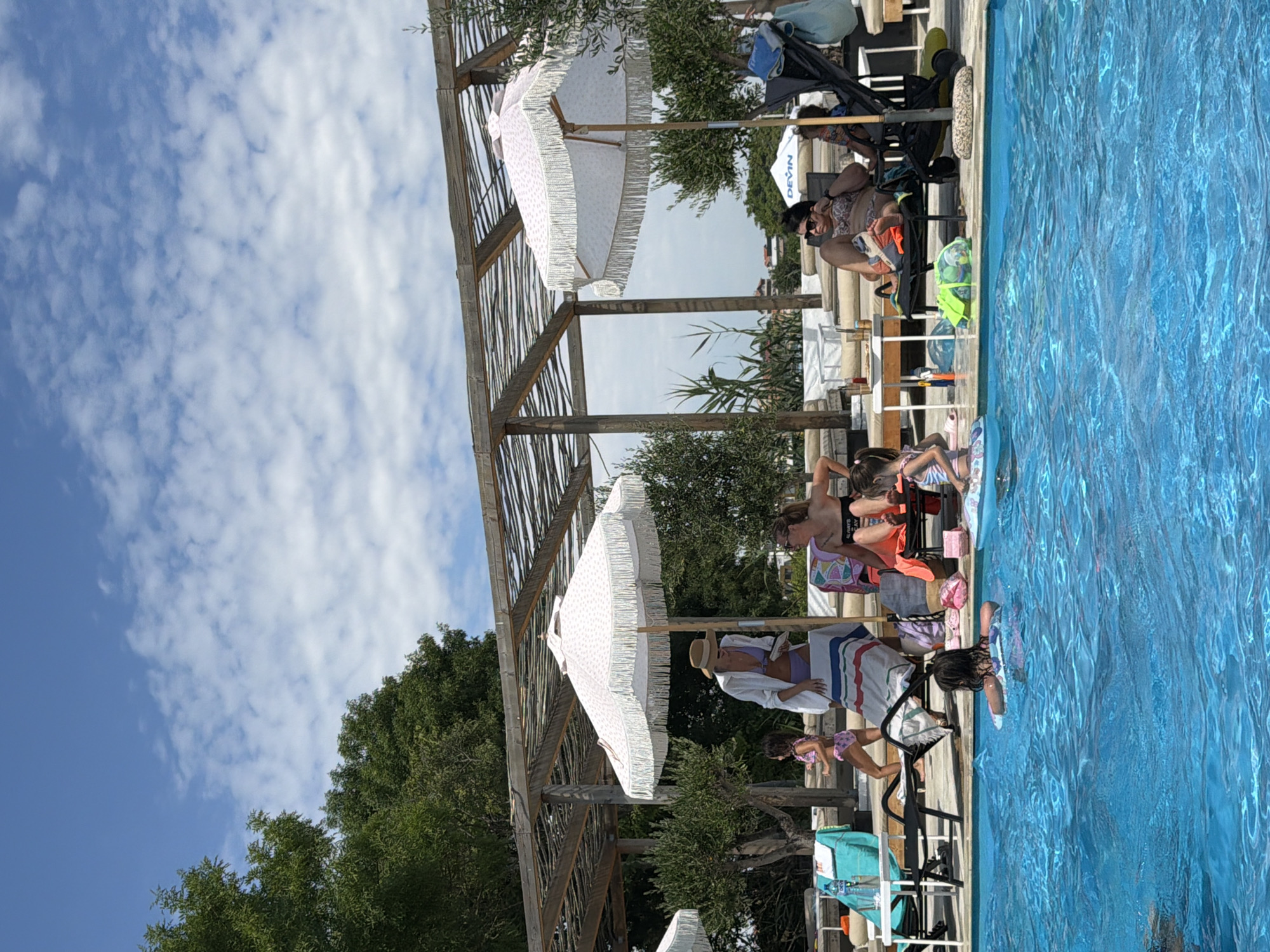 This image depicts a poolside scene at the "Aqua Park" in Burgas, Bulgaria, on a sunny day with a partly cloudy sky. Several people are present, enjoying the water and the amenities.
In the foreground, the bright blue water of the swimming pool occupies the lower half of the image. Several children are visible in the pool. One child, facing away from the camera, is floating on an inflatable ring. Another child, also facing away, is near the edge of the pool on an inflatable mat.
Along the edge of the pool, a white pergola structure provides shade. Underneath the pergola, several lounge chairs are arranged. A woman in a wide-brimmed hat and a white cover-up over a purple bikini is standing near a lounge chair, holding a book. A young girl in a pink swimsuit is standing to her left, near the pool.
Further to the right, a woman in a patterned bikini is seated on a lounge chair, looking towards the right. Next to her, a person is partially visible in a stroller. A second large white umbrella with pink fringe is positioned over this seating area.
Other elements visible include trees with green foliage, lounge chairs with colorful cushions, towels, and beach bags. The overall atmosphere is one of leisure and recreation.