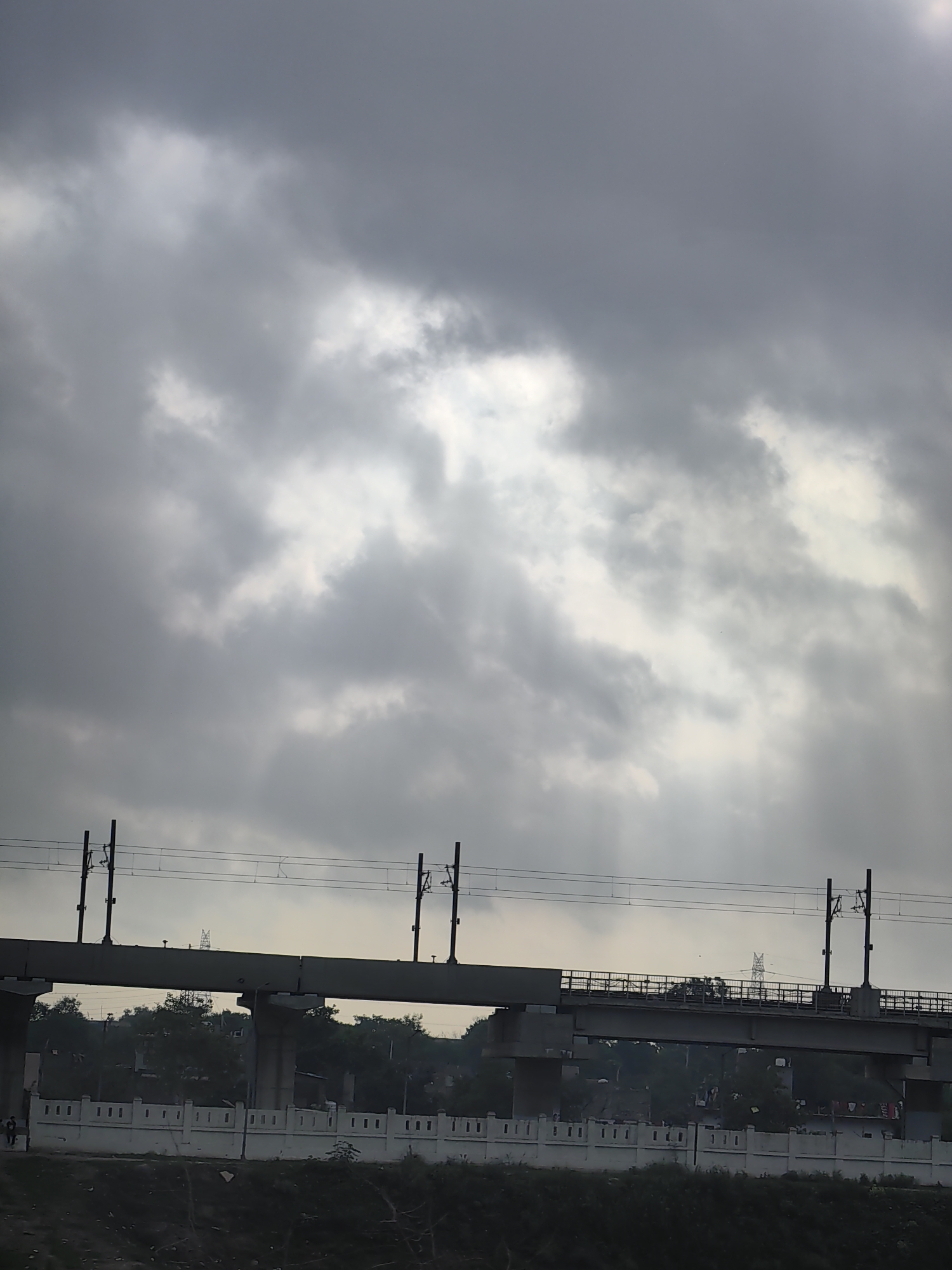 This image depicts an overpass or flyover in Delhi, India, likely part of the Delhi Metro or a similar elevated transit system, viewed from below. The sky is overcast with dramatic, light-penetrating clouds, suggesting an impending storm or recent rainfall. The structure is made of concrete and metal, with visible support pillars and overhead power lines for the train. Below the overpass, there are trees and some indistinct buildings, along with a white, low wall with a decorative pattern, possibly bordering a road or walkway. Two figures are visible near the left side of the frame, appearing to be walking. The overall impression is one of urban infrastructure under a moody sky.