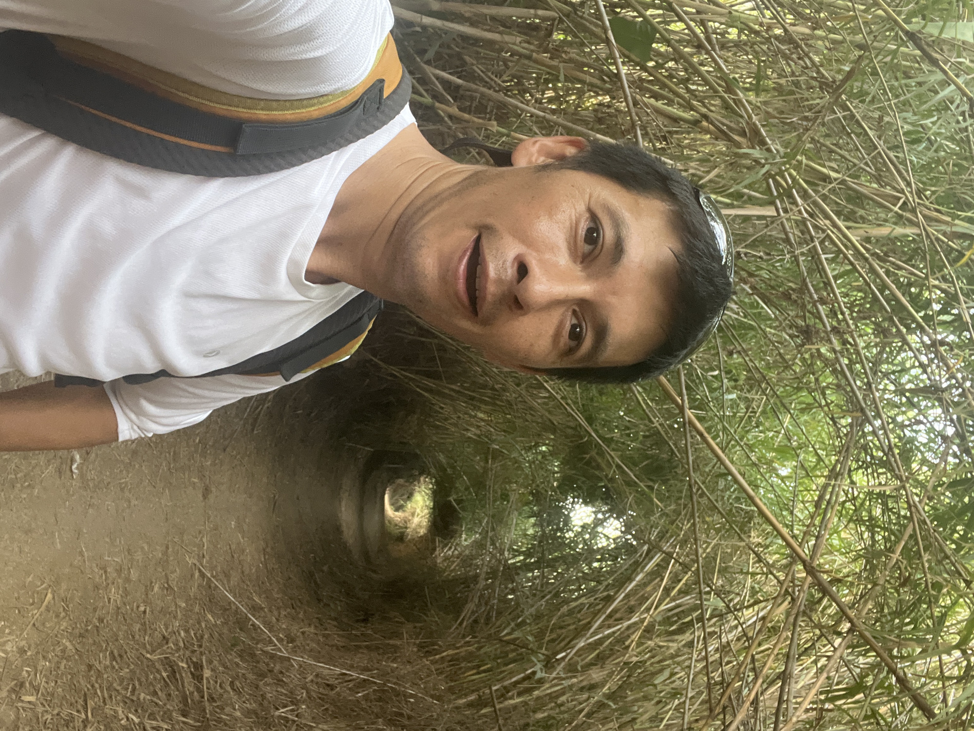The image is a selfie of an adult Asian male wearing a white t-shirt and a backpack. He is standing on a dirt path that is surrounded by dense bamboo growth, forming a natural tunnel. The bamboo stalks arch over the path, creating a shaded passage. The man is looking directly at the camera with a slight smile. The background shows the continuation of the bamboo tunnel receding into the distance, with sunlight filtering through the leaves at the far end. The setting appears to be a natural environment, likely a park or a trail. The location is not identifiable as Tarragona, Spain, based on the visual information provided.