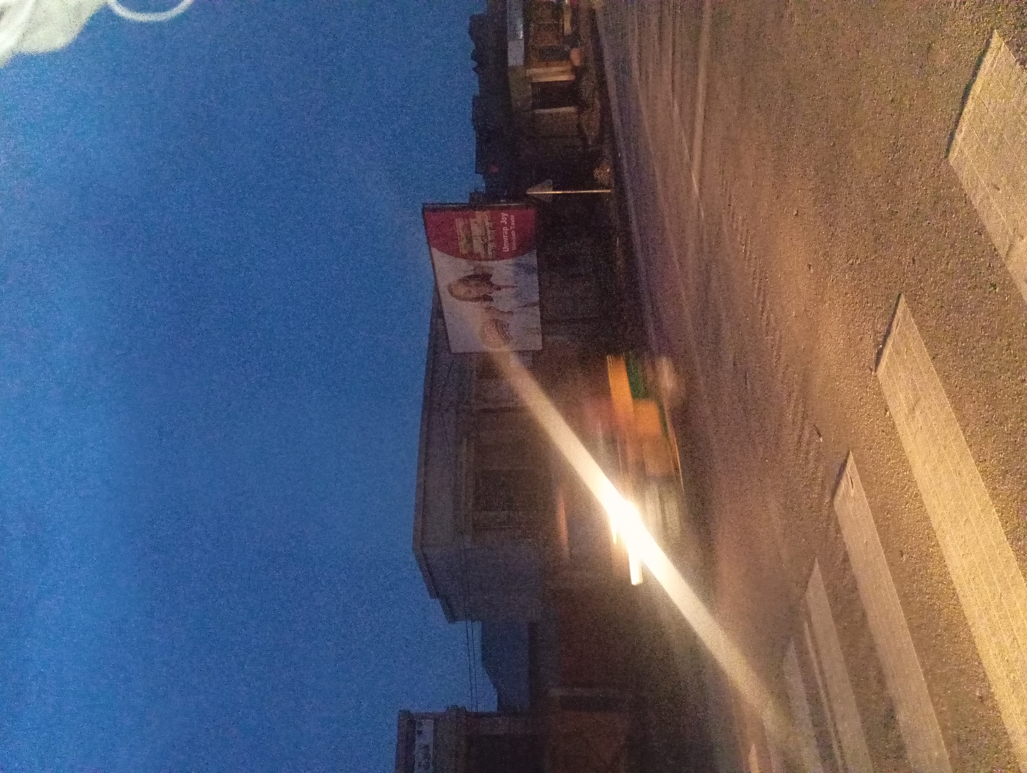 The image captures a street scene at dusk in an urban environment. In the foreground, a pedestrian crossing with white stripes is visible on an asphalt road. To the left, a blurry vehicle with bright headlights is moving through the scene, indicated by a streak of light. In the background, buildings are dimly lit, and a billboard is prominently displayed. The billboard features a woman smiling and text that reads "Unwrap Joy. Ultimate Taste." The sky is a deep blue, transitioning from daylight to night. The overall impression is that of a typical street corner in a city, possibly during twilight hours, with traffic and advertising present.