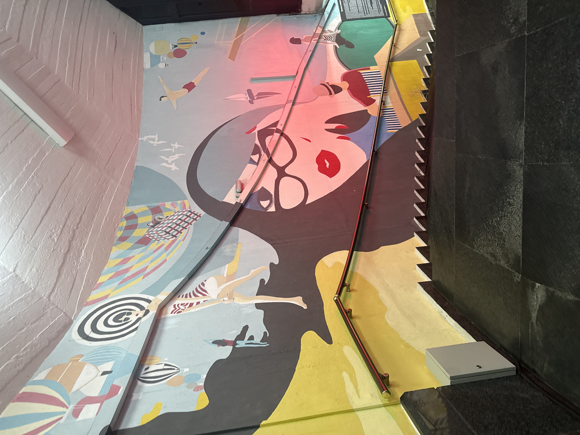 The image captures a mural on a curved wall above a set of stairs. The mural features a stylized, close-up depiction of a woman's face with dark hair, glasses, and red lips. Surrounding the face are various graphic elements, including hot air balloons, geometric patterns, and silhouetted figures, some of whom appear to be swimming or diving. The colors are bold and vibrant, with a pastel blue background. A red light illuminates part of the mural. The stairs themselves are made of dark stone, with metal railings. A light-colored utility box is mounted on the wall to the left of the stairs. The scene appears to be an interior space, possibly a subway station or underpass, given the architectural features. The location is identified as Yalta, Ukraine.
