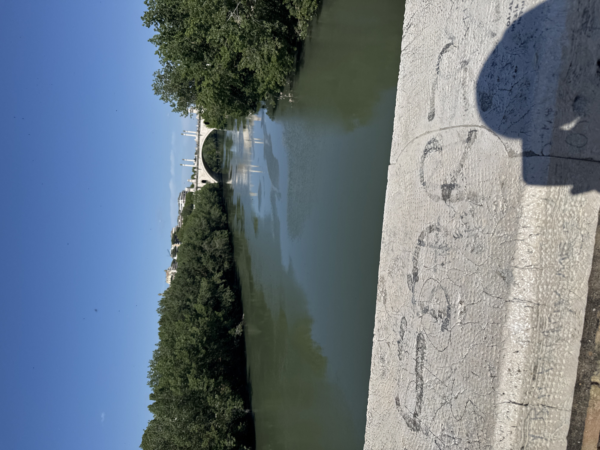 A photograph taken in Rome, Italy, shows the Tiber River flanked by trees. An arched bridge spans the river in the distance. Graffiti is visible on the stone railing in the foreground, with a shadow cast on it. The sky is clear and blue.