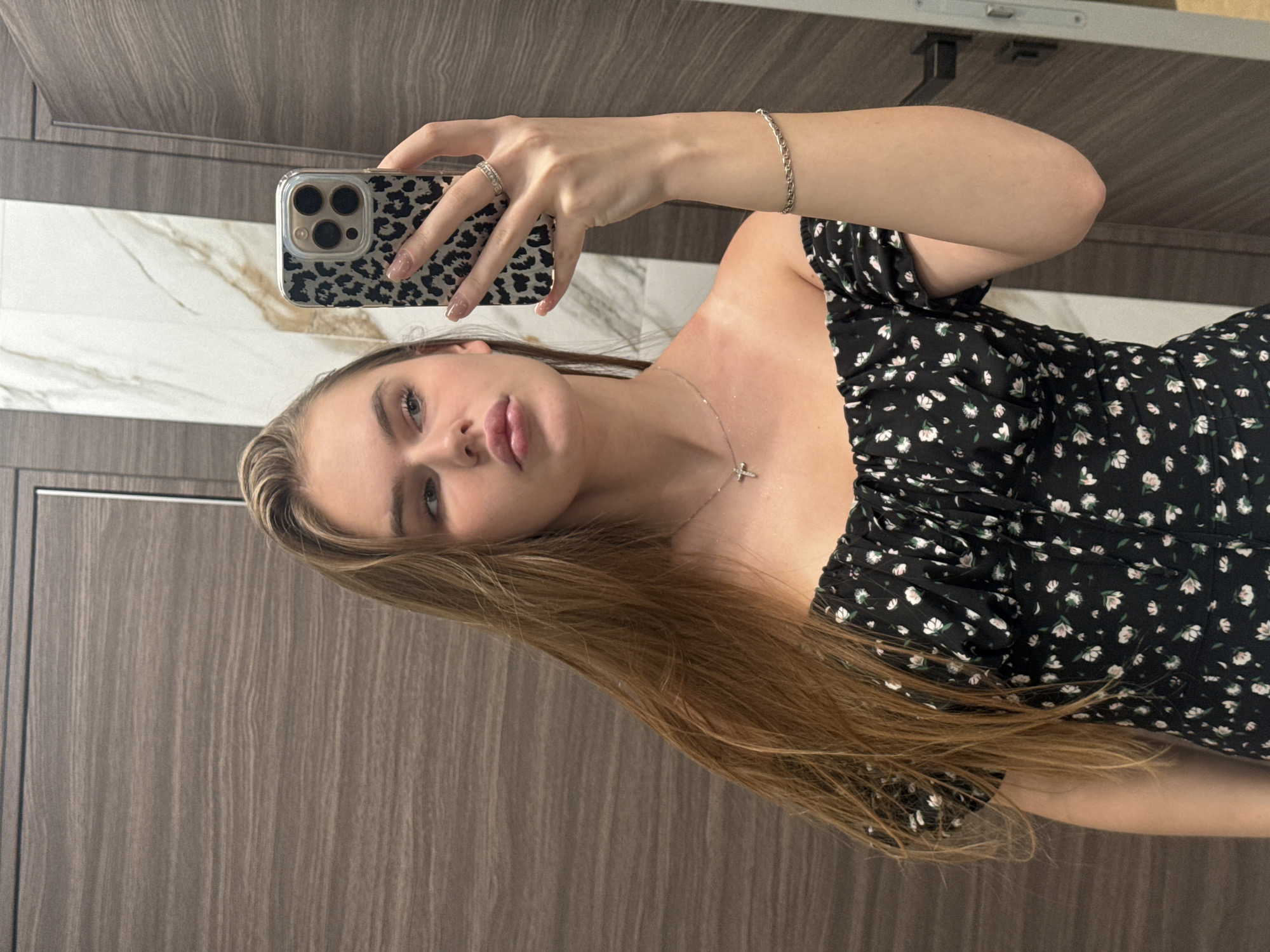 A young woman with long, blonde hair is taking a mirror selfie in a bathroom. She is wearing an off-the-shoulder black dress with a floral print. She has a delicate necklace with a cross pendant and a bracelet on her left wrist, and rings on her right hand. The bathroom features dark wood-paneled walls and a marble accent wall. The overall impression is one of a modern, well-maintained private space. The image does not contain information that can be used to determine the location within the city of Dnipro, Ukraine.