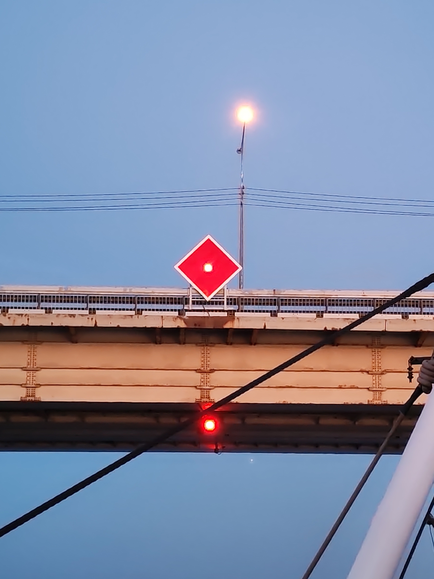 The image shows a bridge structure with a red diamond-shaped sign with a white dot in the center. Below the sign, on a lower section of the bridge, is another red light. Above the bridge, there is a lamppost. There are cables or wires running across the top of the image. The sky is a gradient of blues. The bridge appears to be made of metal, with visible rivets and supports. Part of a pole and rigging is in the lower right corner, suggesting the photo was taken from a boat. The location and city are unknown.