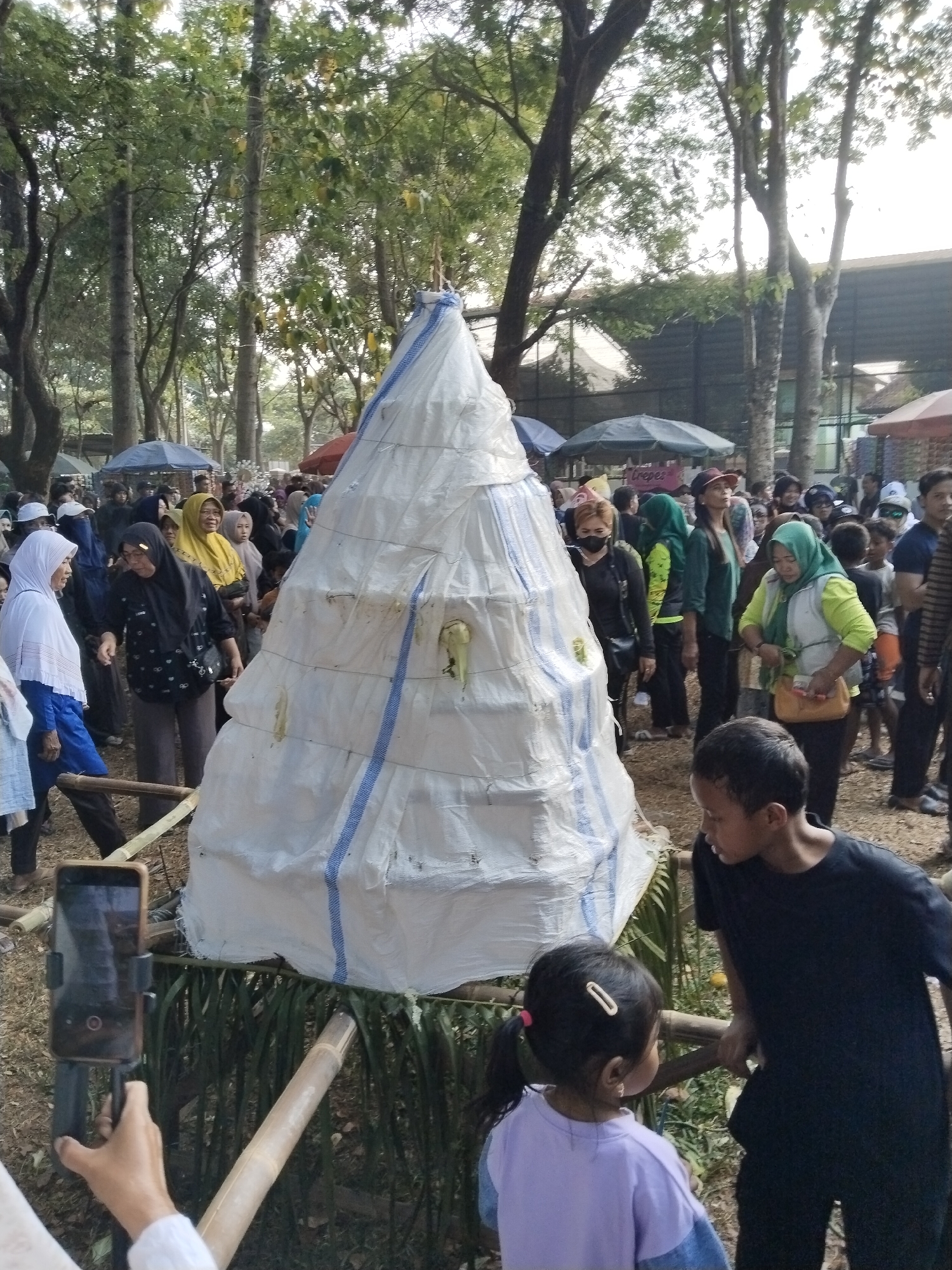 In Karangmalang, Indonesia, a conical structure, possibly a food display or decoration, stands prominently in an outdoor setting, possibly a park or festival grounds. The structure is covered in white material with blue stripes and appears to be made of stacked layers. A variety of people, many of whom are wearing headscarves, are gathered around the structure and in the background. Some individuals are observing the conical object, while others appear to be interacting or engaged in other activities. The environment includes trees with visible foliage, suggesting an outdoor natural setting. In the foreground, a person is holding a smartphone on a tripod, likely recording the event. Two children are also visible in the foreground, one looking towards the other who is leaning on a wooden railing. The overall scene suggests a community gathering or celebration.