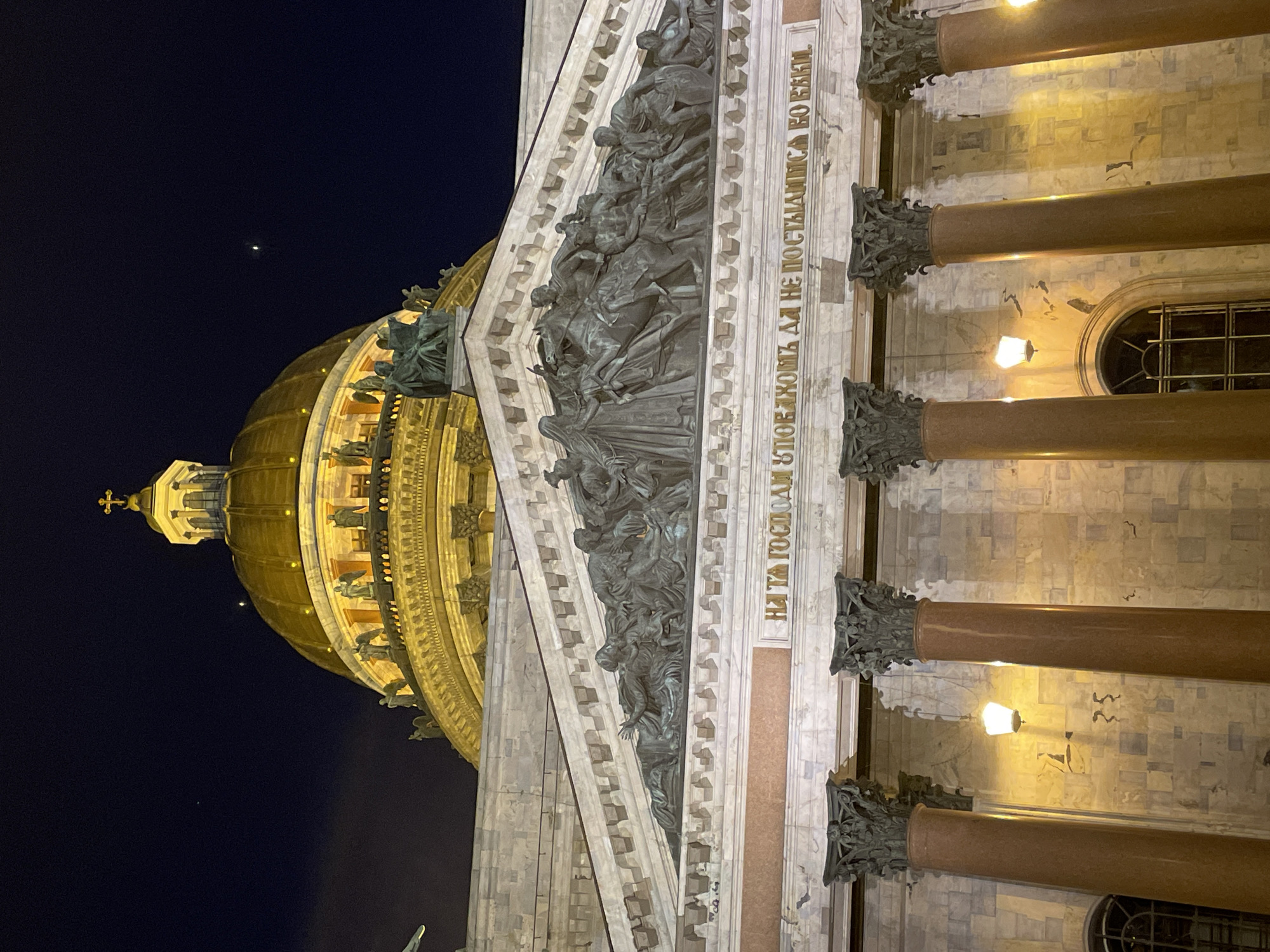 A nighttime, vertical shot captures a grand architectural edifice, likely a cathedral or public building, with a prominent gilded dome against a dark sky. The structure is illuminated by artificial lights, highlighting its details and creating a warm glow.
The main subject is a large, classical-style building facade featuring massive columns with ornate capitals. Behind and above these columns, a wide frieze displays intricate bronze bas-relief sculptures depicting numerous figures in dynamic poses, including horses and individuals in dramatic scenes. Above this, a golden dome rises, topped with a smaller cupola and a cross. Statues are visible around the base of the dome and on the lower parts of the building. Windows, some with grates, are visible on the building's stone walls.
The time of day is clearly night, with the sky appearing very dark blue or black. A single bright celestial object, possibly a star or planet, is visible in the upper sky to the left of the dome. The artificial lighting on the building creates strong contrasts and emphasizes the architectural and sculptural details.
Visible text in Cyrillic is inscribed on a frieze above the columns: "НА ТА ГОСПОДАМ А ДА НЕ ПОСТЫЛНТСА ВО БЕКИИ." This phrase is an old Church Slavonic inscription, likely a biblical quote (e.g., from Psalms), which roughly translates to "In Thee, O Lord, have I trusted; let me never be put to shame forever." The solemnity and grandeur of the architecture, combined with the illuminated sculptures and text, evoke a sense of reverence and historical significance.