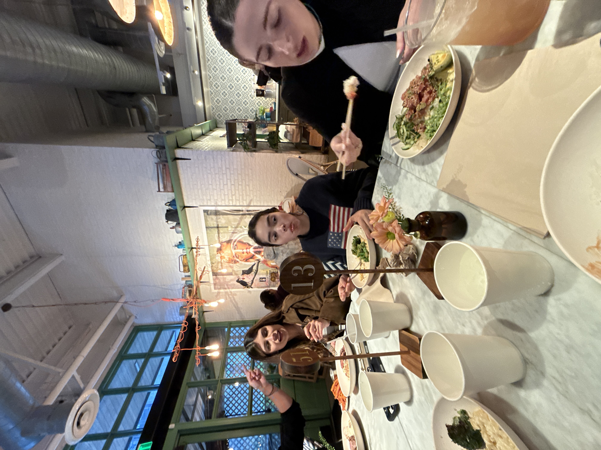 Captured indoors in the city of Austin, United States of America, the scene unfolds in what appears to be a restaurant with an indoor setting. The setting showcases a group of individuals gathered around a table, engaged in dining activities. People are seen eating and interacting with each other, contributing to the dynamic of a shared meal.
The dining table is arranged with plates filled with food, along with glasses and utensils, indicative of a meal in progress. A wooden table number prop adds to the restaurant ambiance.
The restaurant has a distinctive decor with exposed ductwork on the ceiling. Large windows with a green frame provide natural light, adding to the atmosphere. The restaurant is further decorated with hanging lights.