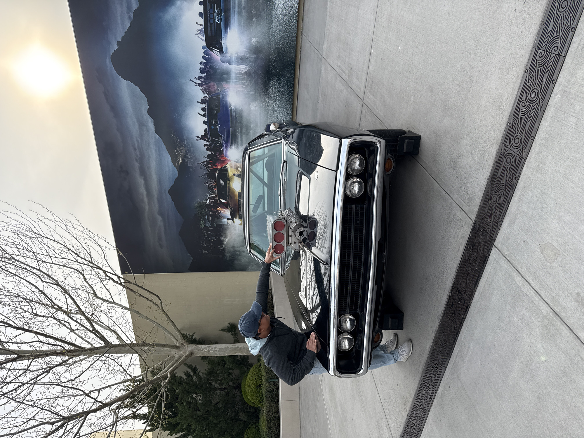 A man in a black jacket, light blue hoodie, blue jeans, and gray sneakers, is touching the hood of a black car. The car has a silver grill, chrome front bumper, and a large chrome engine modification protruding from the hood. Behind the car is a large mural of a movie scene. To the left of the mural is a building wall and a bare tree. To the right of the car is a gray concrete sidewalk with a decorative black border. The photograph was most likely taken during the day, outdoors. There are no clear identifiers that indicate the location is Tongzhou District, China.