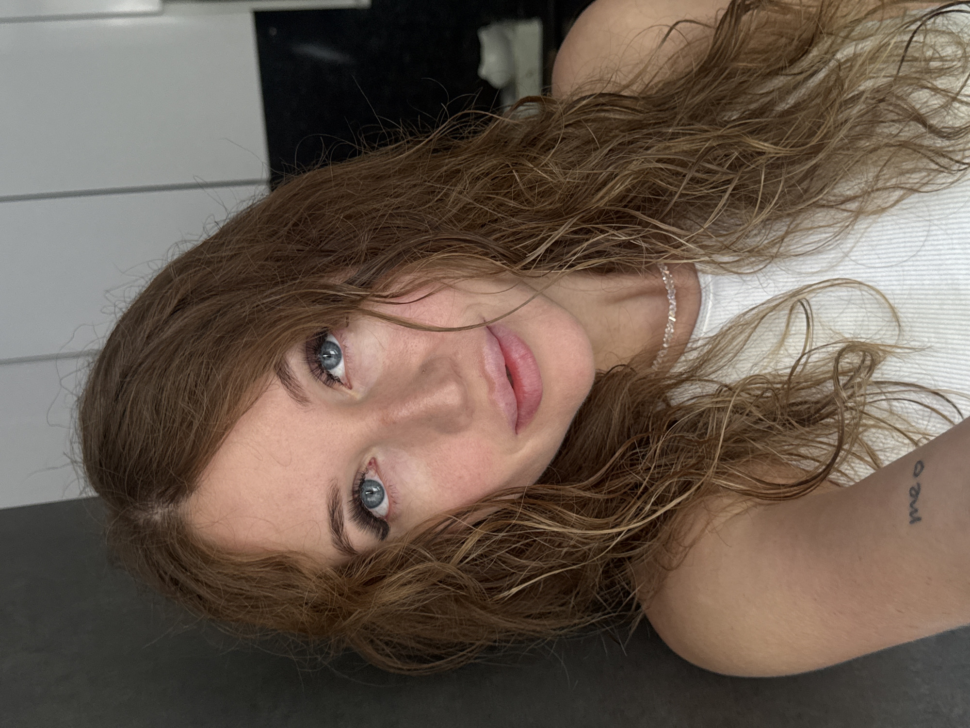 A close-up, head-and-shoulders selfie of a young Caucasian woman with light brown, wavy hair and blue eyes. She is wearing a white, ribbed tank top and a delicate beaded necklace. Her left arm is visible, showing a small tattoo on her inner bicep that appears to read "meow." She is leaning against a dark gray wall, and the background behind her is a softly blurred kitchen interior with white cabinets and black countertops. There is no indication of the city of Chaikovsky, Russia.