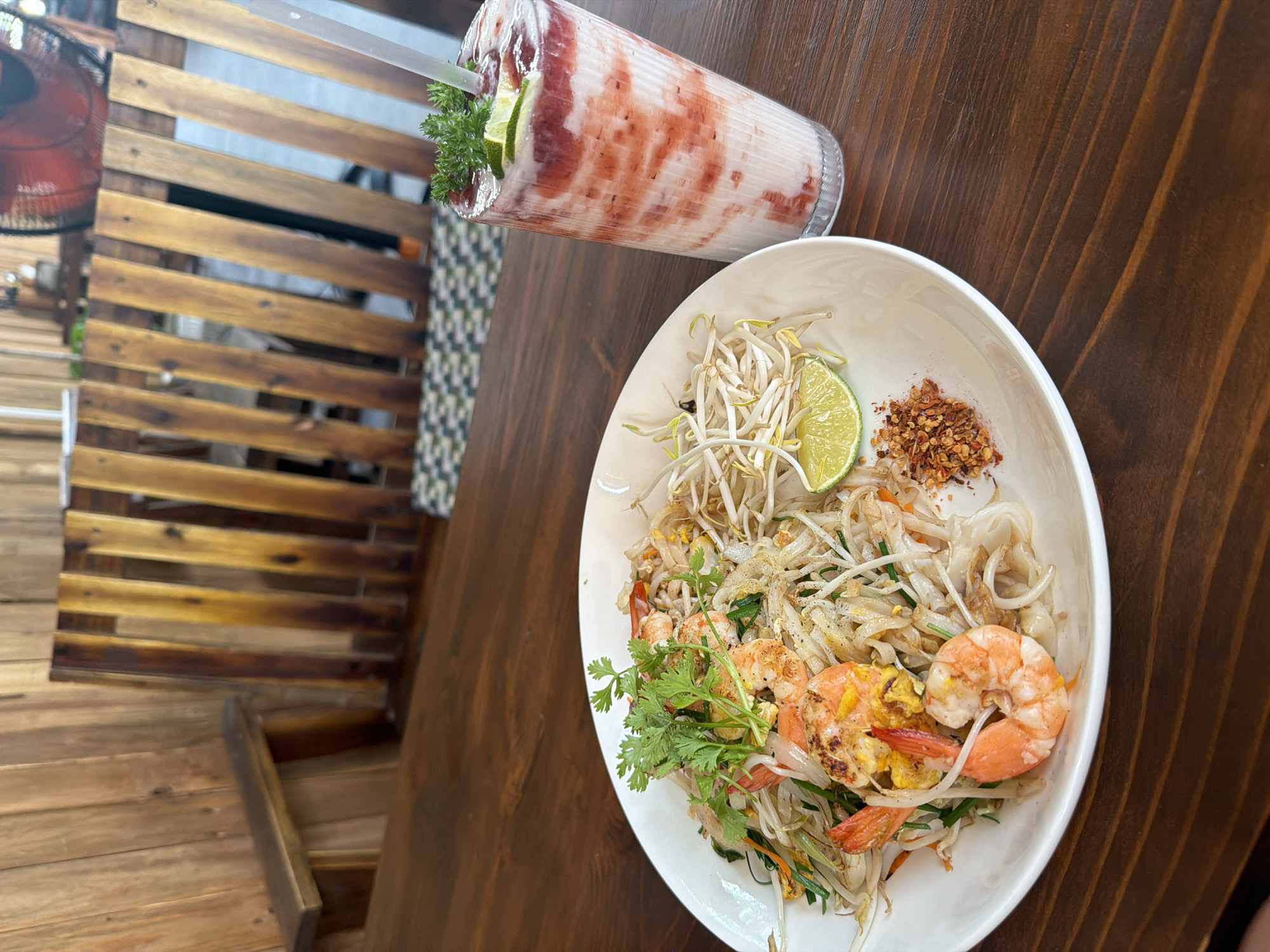 A partially-filled white bowl containing Pad Thai noodles with shrimp, bean sprouts, cilantro, a lime wedge, and crushed red pepper flakes sits on a dark brown wooden table. Adjacent to the bowl is a tall glass containing a pink and white layered beverage garnished with a sprig of herbs and a lime wedge. The background includes a partially visible red electric fan, a patterned screen, and a wooden wall. The location is Nha Trang Ward, Vietnam, indicated by the food and environment.