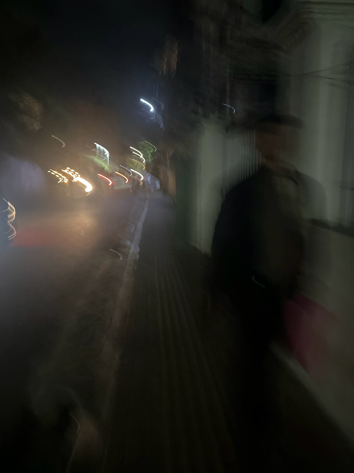 This is a photograph taken at night in the city of Lalitpur, Nepal. The image depicts a blurry, abstract scene with streaks of light, suggesting movement during the exposure. In the foreground, there are out-of-focus yellow and white streaks of light, possibly from passing vehicles or streetlights. To the right, a dark, indistinct figure of a person is visible, also blurred by motion. The person appears to be wearing a jacket and a hat. The background shows dark shapes of buildings or trees and a lighter, vertical surface that could be a wall. The overall impression is one of a dynamic urban environment at night, captured with a sense of motion and low light.
