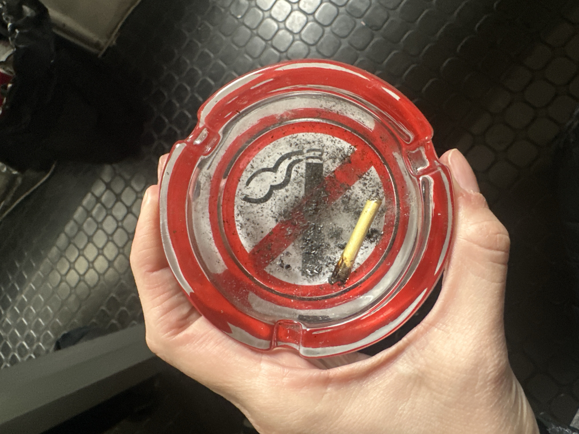A hand holds a round, clear glass ashtray with a red border. The ashtray contains ash and a spent cigarette. The ashtray features a "no smoking" symbol. The background is blurry. I cannot determine the location in Buenos Aires, Argentina, or any other city, based on the image.