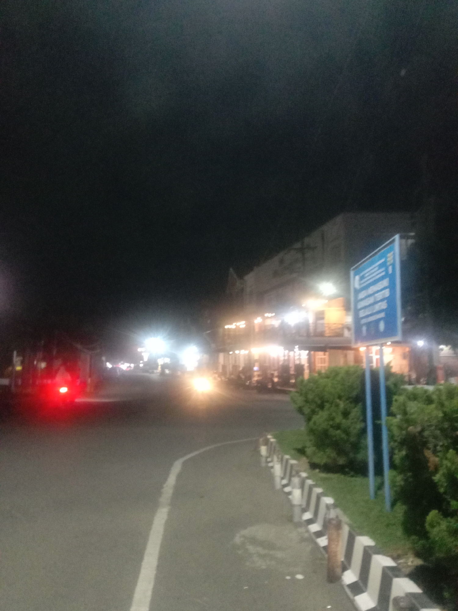 The image depicts a blurred, low-light nighttime scene on a street, likely in an urban or semi-urban area. The time of day is dark night, with artificial lights serving as the primary illumination.
In the foreground, a paved road with a single solid white line marking a lane is visible. On the left side of the road, a bright red taillight from a vehicle (possibly a motorcycle or car) is prominent and blurred. Further into the distance on the left, other indistinct lights suggest additional traffic or distant structures. On the right side of the road, a bright, yellow-white headlight from an approaching vehicle creates a strong light source.
To the immediate right of the road, there's a patterned curb or barrier featuring distinct black and white diagonal stripes. Beyond this, a patch of green grass and several green bushes or shrubs are visible, followed by a tall, blue informational sign mounted on blue poles. The sign has white text, with several lines clearly legible in Indonesian: "KEMENTERIAN KESEHATAN REPUBLIK INDONESIA" (Ministry of Health Republic of Indonesia) at the top, followed by "BADAN LAYANAN UMUM PUSKESMAS" (Public Service Agency Community Health Center). Further text is present below but is too blurry to decipher.
Behind the sign and vegetation, a multi-story building is visible, with its lower floors brightly illuminated, suggesting commercial activity such as shops or restaurants. The upper floors are darker and less distinct. The overall impression is one of a bustling street scene at night, characterized by motion blur and vibrant artificial lights against a dark backdrop. No specific individuals are discernible, but the presence of vehicles and lit establishments implies human activity. The general setting appears to be a roadside area within a town or city, consistent with Lhokseumawe, Indonesia.