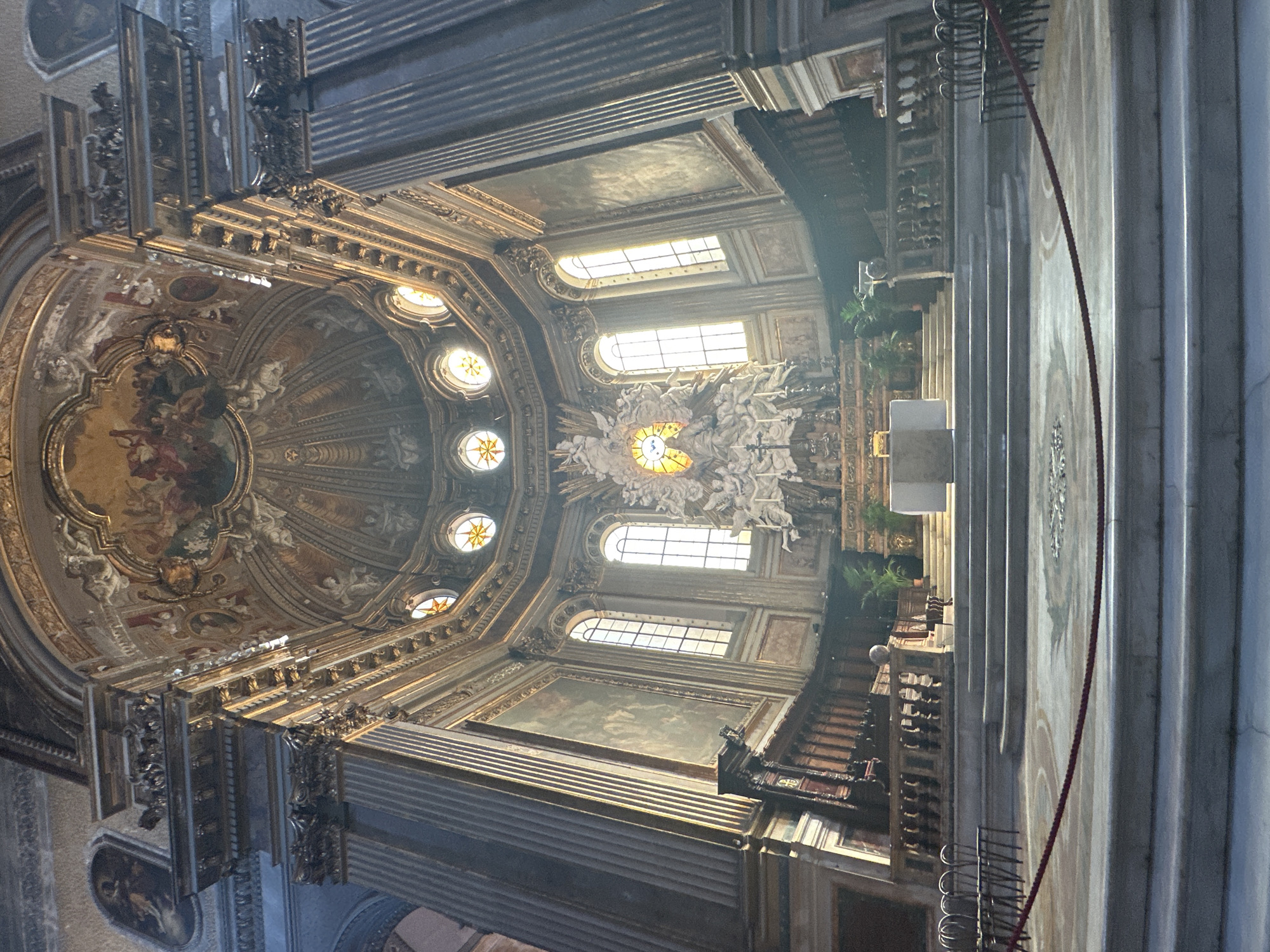 The image shows the interior of the church of Gesù Nuovo in Naples, Italy. It is a wide-angle shot looking towards the altar. The church features a grand baroque interior with a large dome adorned with frescoes. There is an elaborate altar with statues and stained glass windows. In the foreground, there are several levels of marble steps leading up to the altar area. Rows of empty chairs are visible on either side. Sunlight streams in through the windows, illuminating parts of the church and creating dramatic shadows. The overall impression is one of grandeur and solemnity.