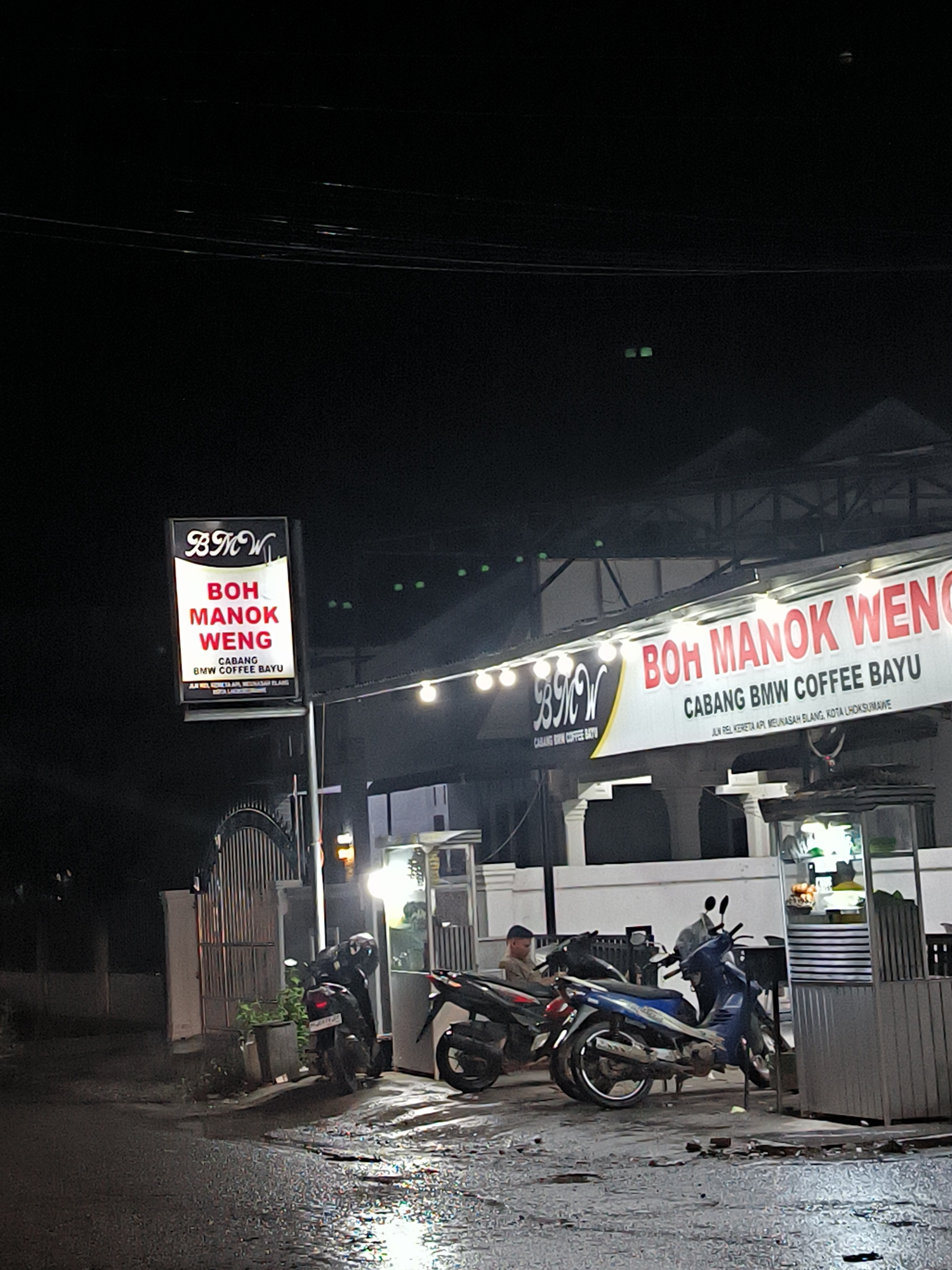 The image captures a nocturnal outdoor scene in Lhokseumawe, Indonesia, depicting a coffee shop or eatery at night. The foreground shows a wet asphalt road, reflecting the artificial lights from the establishment, suggesting recent rain. Several motorcycles are parked on the damp ground in front of the building, with one person sitting on a motorcycle near the entrance.
The main subject is a brightly lit building housing "BOH MANOK WENG CABANG BMW COFFEE BAYU." A prominent vertical sign on the left side of the road also displays "BMW," "BOH MANOK WENG," and "CABANG BMW COFFEE BAYU." Both the banner on the building and the freestanding sign include the address: "JL. REL KERETA API MEUNASAH BLANG, KOTA LHOKSUMAWE." String lights are draped across the front facade of the building, illuminating the area. A small food cart or stall is visible to the right of the entrance. In the dark background, faint green lights are visible, possibly from another distant source or signage. The overall atmosphere is quiet and illuminated by artificial light against the dark sky.