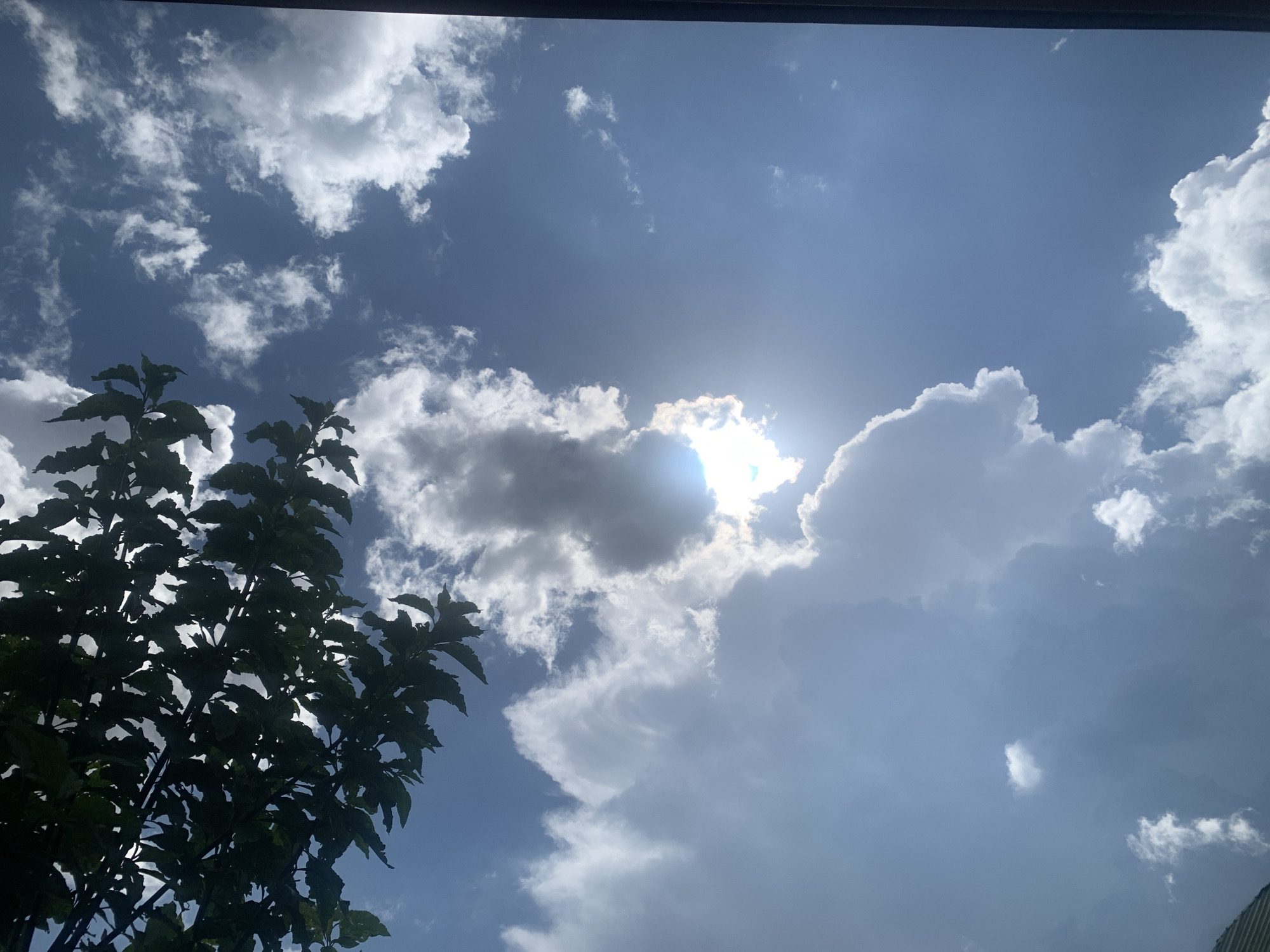 The image depicts a sky with scattered clouds and the sun partially obscured by clouds. A portion of a dark green leafy tree is visible in the lower left quadrant of the image. The scene appears to be outdoors, with a view of the sky from a ground-level perspective, possibly from a window or beneath a structure, indicated by a dark horizontal bar across the top. The lighting suggests it is daytime, with the sun casting light and creating some glare. The location is identified as Kuru Karama, Nigeria. There are no people or specific objects of interest, nor any identifiable actions or interactions taking place.
