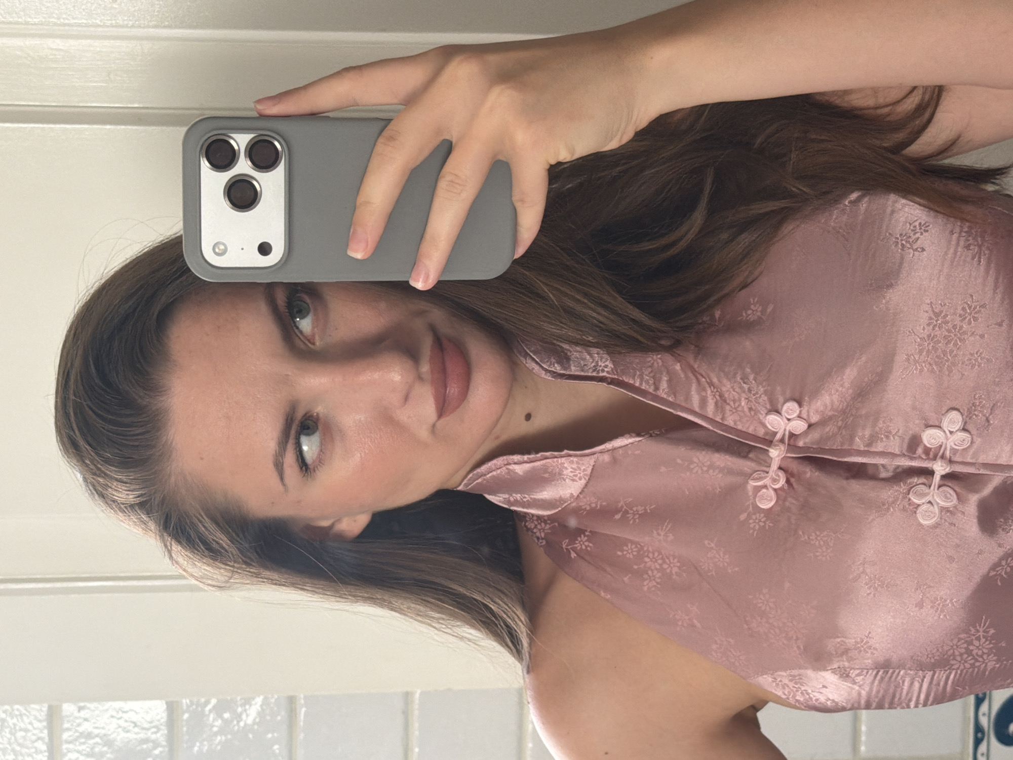 A young woman is taking a selfie in a bathroom. She has long, brown hair and is wearing a pink, satin halter top with a floral pattern and frog closures. She is holding up a grey smartphone to take the picture. The background appears to be a tiled wall and a door. There is no indication of the image being taken in Sharm El Sheikh, Egypt.