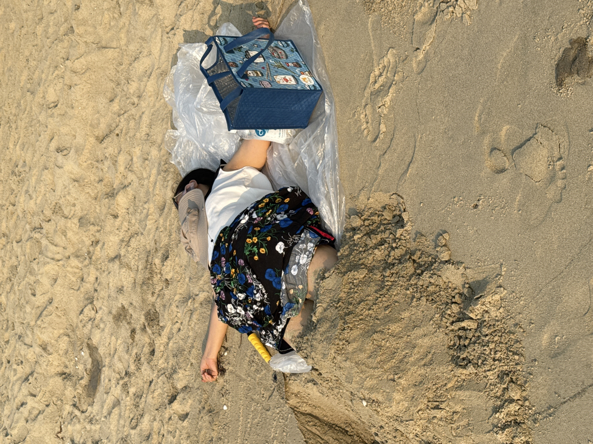 The image depicts a person lying on the sand at a beach in Đà Nẵng, Vietnam. The individual is partially lying on a clear plastic sheet with a blue, patterned insulated cooler bag resting on their legs. They are wearing a white shirt, a floral print skirt, and a face mask. A tan-colored cardigan or jacket is visible. A yellow-handled trowel encased in a clear plastic bag is located near their legs. The sand varies in color and texture, with footprints visible in one section.