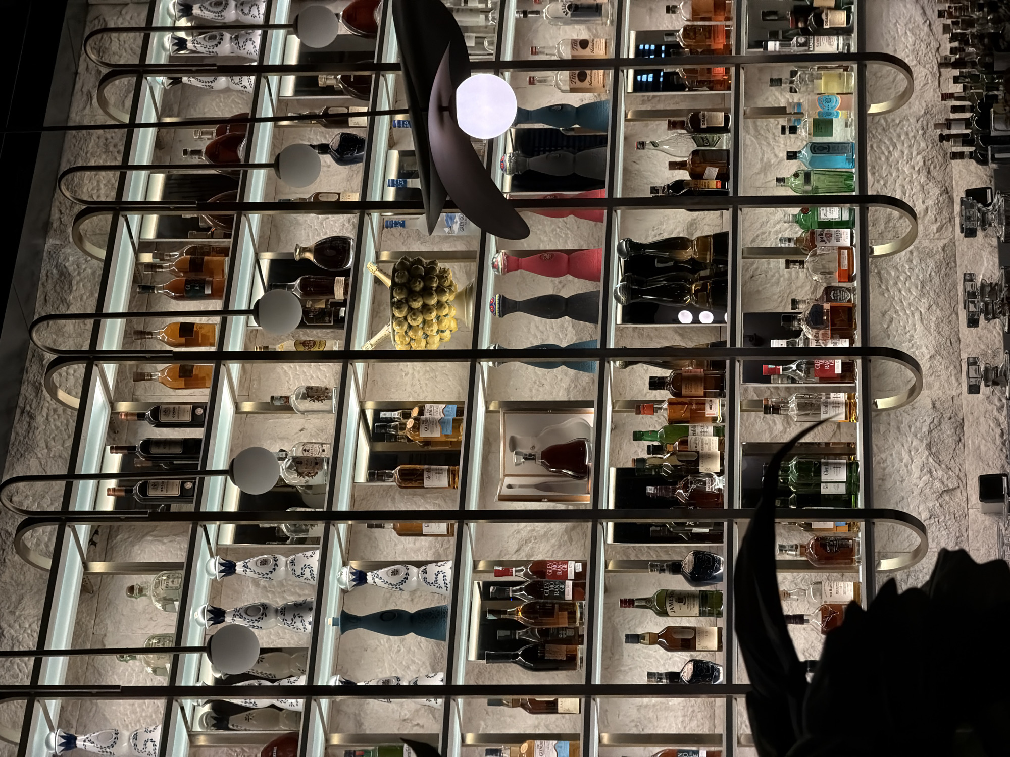 The image shows a detailed view of a backlit, grid-like alcohol display, featuring numerous bottles arranged on shelves. The bottles are a mix of different shapes, sizes, and brands, suggesting a diverse selection of spirits. The display is illuminated from behind, creating a soft glow that highlights the bottles and the textured wall behind it. A decorative lamp with a black, curvilinear design and a white globe light is prominent in the foreground, slightly obscuring some of the bottles. The overall composition is a mix of geometric order in the shelving and a more organic feel from the variety of bottles and the decorative lamp. There is also an object in the right side of the frame in the bottom corner that is out of focus, which appears to be a statue, most likely a dragon. Some of the bottles are recognizable as Jameson and Glenlivet. The surrounding location is in the city of Budva, Montenegro.