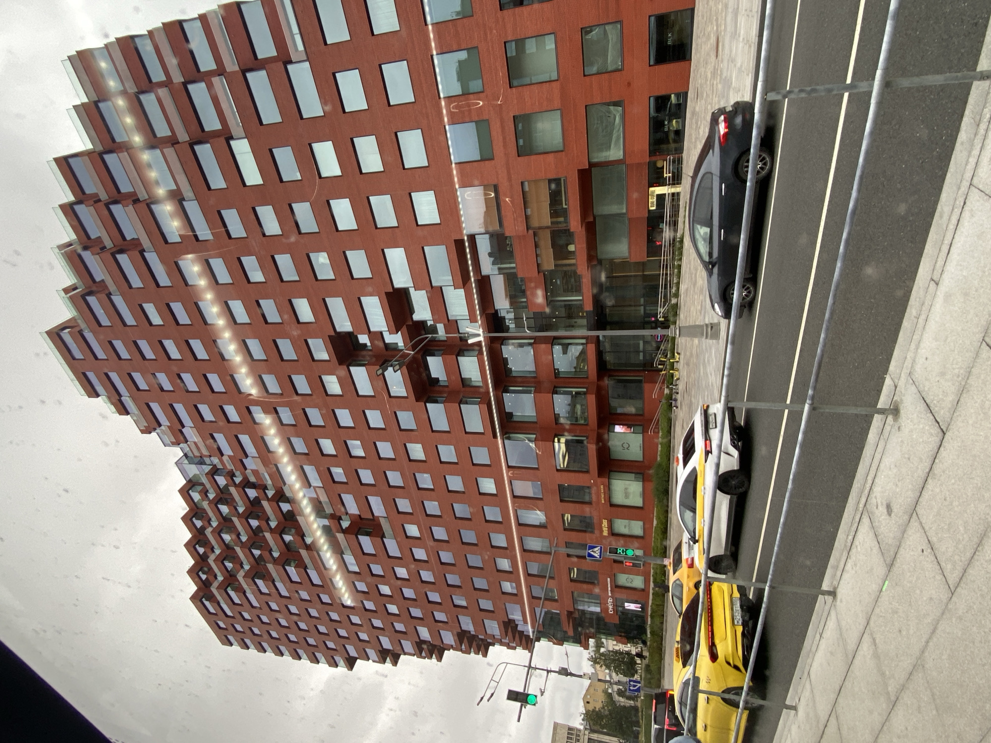 The image captures a street-level view of a modern, multi-story building with a distinctive red brick facade and staggered, cantilevered balconies. The building's architecture is characterized by its repetitive rectangular window patterns. In the foreground, several vehicles are visible on a street, including a yellow taxi, a white taxi, and a dark-colored sedan. A traffic light displaying a green signal is positioned on the left side of the image. The scene appears to be in an urban environment, likely Moscow, Russia, given the presence of Russian license plates and Cyrillic signage on the building's ground floor, which includes text like "ЮЖУУЛЬ" and "WORLD CLASS". The sky is overcast, suggesting a cloudy day. A pedestrian walkway with a grey tiled surface is present on the left side, separated from the road by a metal barrier.