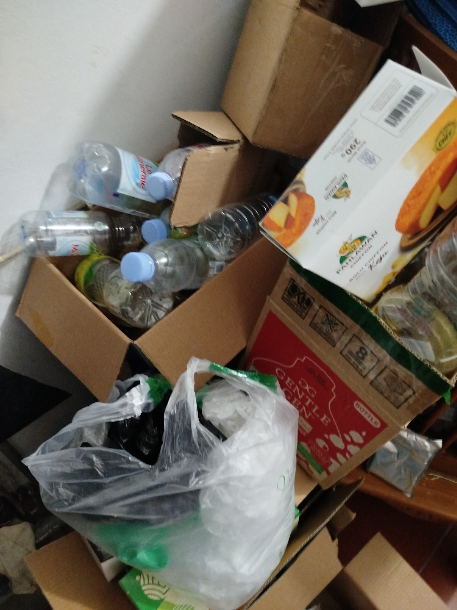 The image depicts a messy pile of recyclables and packaging materials in what appears to be a corner of a room. Cardboard boxes are stacked haphazardly, with some open and others containing various items. Among the items are numerous plastic water bottles, some with blue caps, and a few other plastic containers. A large clear plastic bag, partially filled with dark-colored items and some green material, sits prominently in the foreground, spilling out onto the cardboard. A white cardboard box with an image of cake and text that reads "PAHLAWAN CHIFFON" and "290g" is visible amongst the clutter, leaning against other boxes. Another red and brown cardboard box with text and symbols, including the word "BOTTLE," is also present. The environment suggests a storage area or a collection point for recycling. The lighting is somewhat dim, and the overall impression is one of disarray. The specific location in Bandungrejo, Indonesia, is not evident from the visual information in the image.