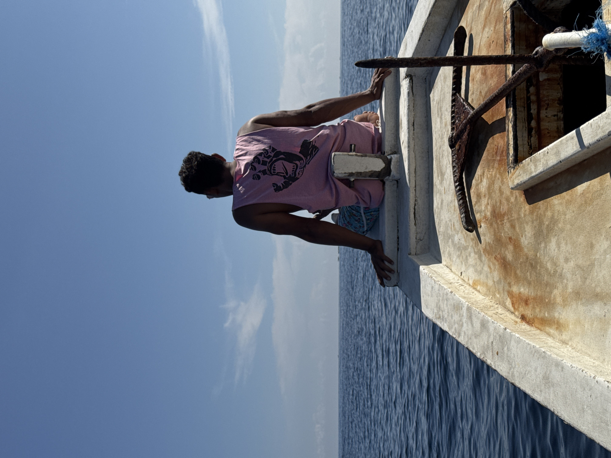 A man is perched on the edge of a boat, possibly fishing, on a clear day in Unknown, Indonesia. The man is wearing a pink tank top with a dark graphic and patterned shorts. He is leaning over the side of the boat, with his hands gripping the edge. An anchor is attached to the side of the weathered boat. The sky is blue with a few wispy clouds, and the sea stretches to the horizon.