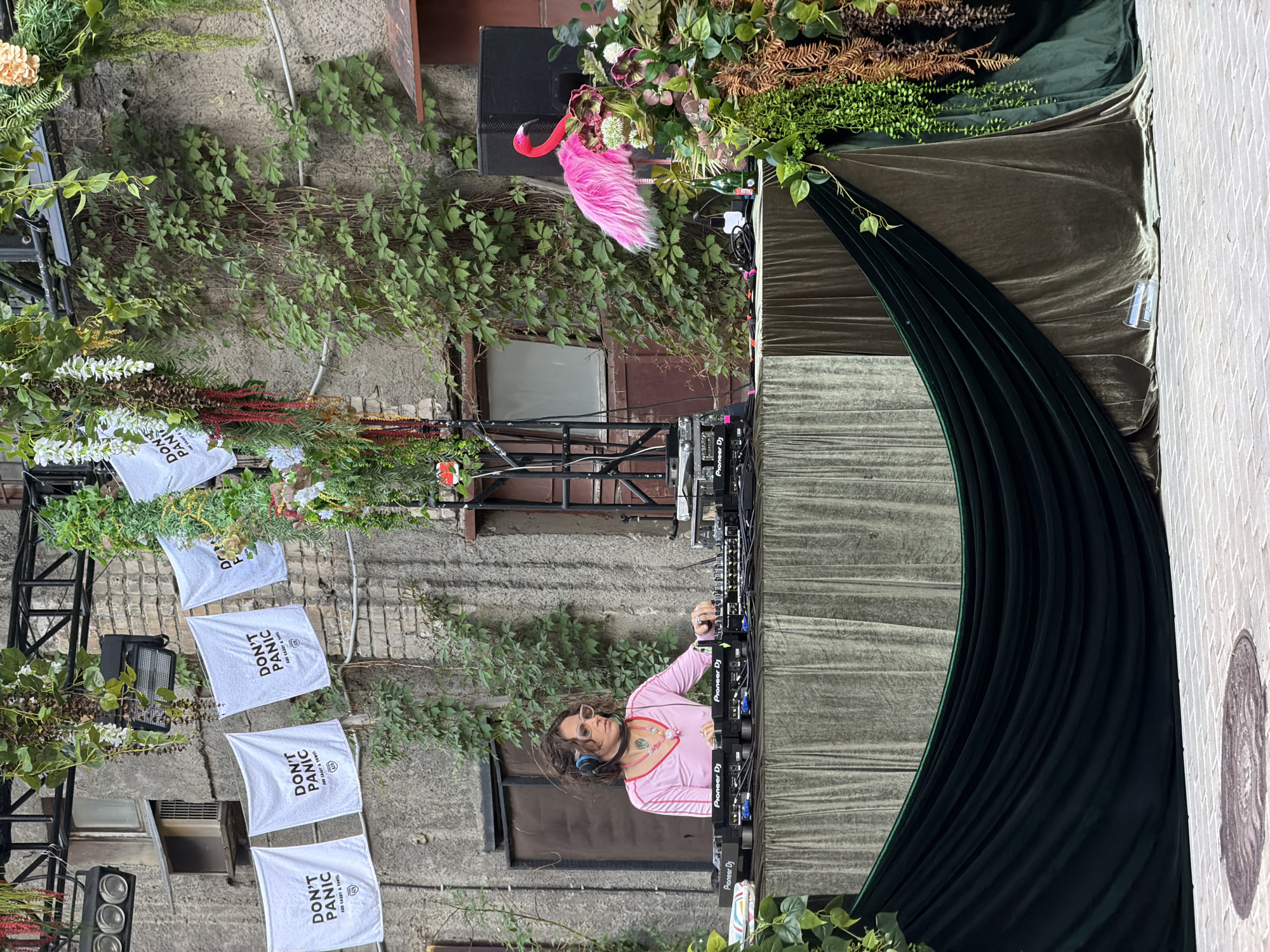 The image captures a DJ performing outdoors against a backdrop of what appears to be an old brick building or wall covered with climbing plants and foliage. Five white towels that read "DON'T PANIC AND CARRY A TOWEL" are tied to a truss-like structure. A DJ console is prominently featured, and a person in a pink top and sunglasses is operating it. Decorative elements, including a pink flamingo and floral arrangements, add to the visual complexity of the setup. The area is decorated with green velvet draping and plant material. There are no clues to confirm the city of Moscow, Russia.