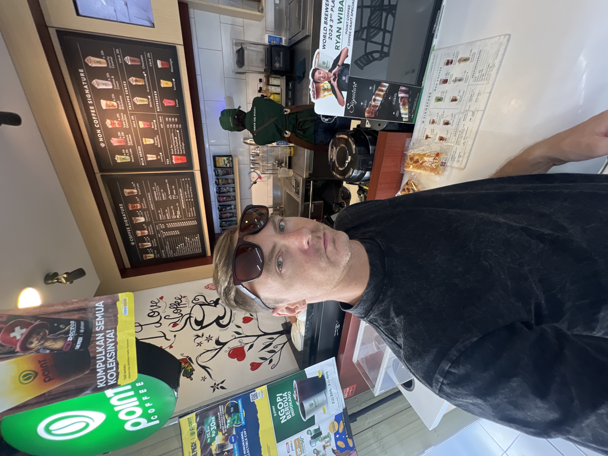 A man wearing sunglasses and a black t-shirt takes a selfie in a Point Coffee shop in Sukawati, Indonesia. He stands in the foreground, with a counter and menu boards behind him. A barista in a green uniform and apron works behind the counter. The shop is decorated with posters and artwork related to coffee.
