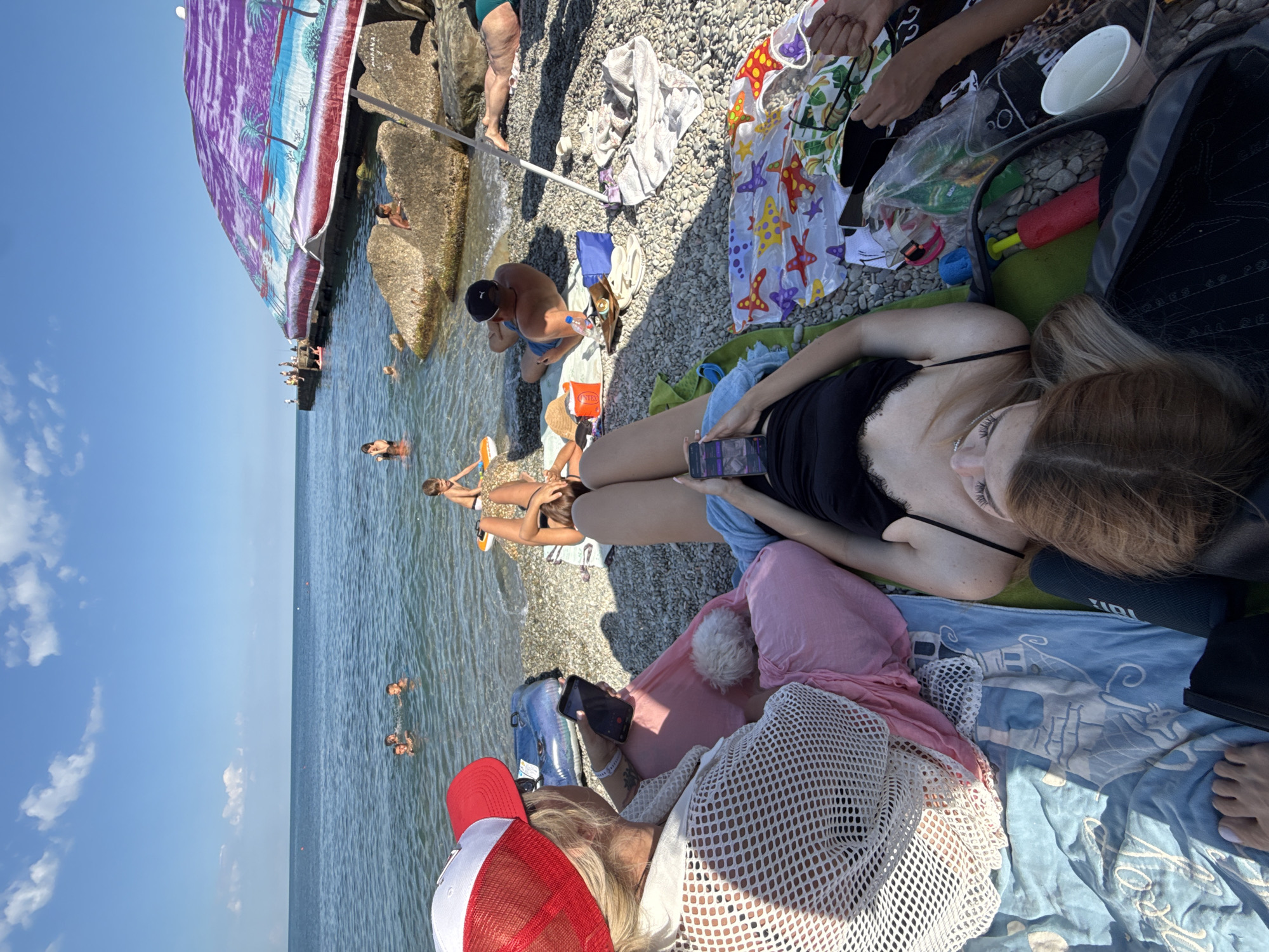 In a wide, overhead shot on a sunlit beach with a pebble shore, a group of people relax near the water. In the foreground, a woman with blonde hair is reclined on a blue towel, holding a smartphone. To her left, another person wearing a red and white cap is also seated, looking at a smartphone. Between them is a pink blanket and a small white dog. Further into the background, several people are in the water, swimming and playing. A large, colorful umbrella with a tropical design is set up to the right, providing shade. The sea extends to the horizon under a bright blue sky with scattered clouds. Rocks and a pier are visible along the coastline. The scene depicts a leisurely day at the beach.