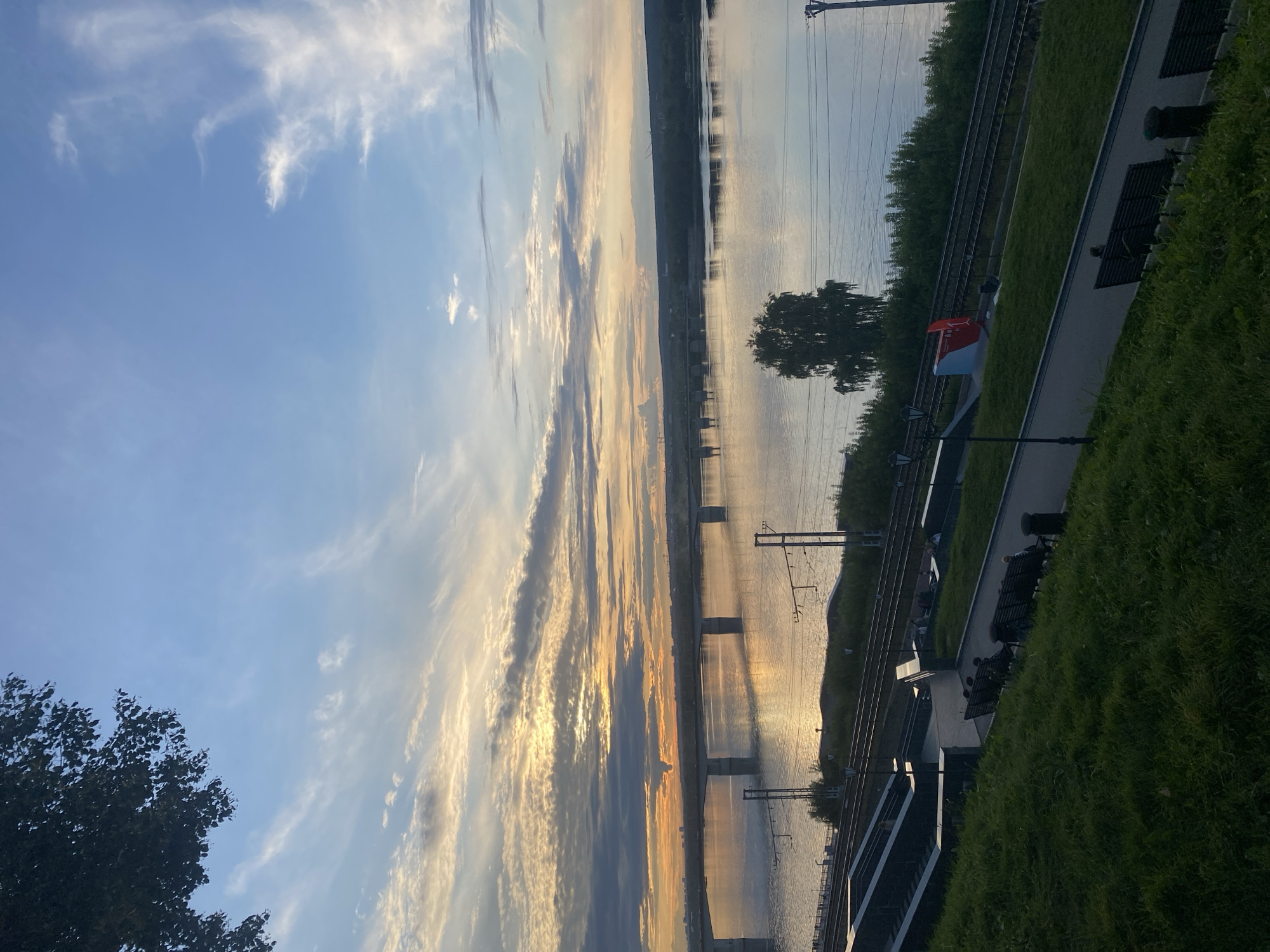 This image depicts a sunset over the Kama River in Perm, Russia, likely from the embankment area near the Monument to the First Teacher.
In the foreground, a grassy slope slopes down towards a paved walkway. Several dark benches are positioned along the walkway, some occupied by individuals. To the left of the walkway, dark stone stairs descend. Further down the slope, a train track runs parallel to the river. Adjacent to the train track, there is a red and blue monument, likely the Monument to the First Teacher. A single tree stands in the midground on the opposite bank of the river.
The midground is dominated by the wide Kama River, reflecting the warm colors of the sunset. A long bridge, supported by concrete piers, spans the river in the distance. The far bank of the river is lined with trees and buildings, with some construction cranes visible on the horizon.
The sky is a vibrant mix of blue, orange, and yellow, with wispy clouds illuminated by the setting sun. The top left corner of the image shows the dark silhouette of tree branches against the sky. The overall impression is one of a peaceful evening scene by the river.