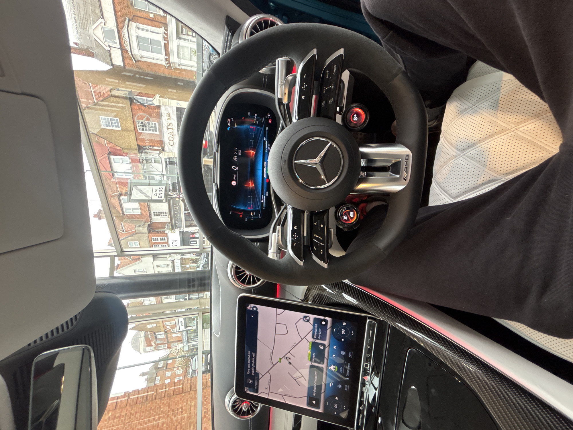 Here is a description of the image:
The image depicts the interior of a Mercedes-AMG car, focusing on the steering wheel and dashboard. A person wearing black trousers is visible in the right side of the frame. The steering wheel is black and features the Mercedes logo, as well as various controls and buttons. Behind the steering wheel is a digital display. The dashboard includes a large touchscreen displaying a navigation map. Outside the car window, a reflection of the street scene is visible, including a building facade with "COATS & Co" signage, street signage "Zone Ends" and "PIACERE" as well as "CONSIGNE DE RODA" suggesting a location in London, United Kingdom.