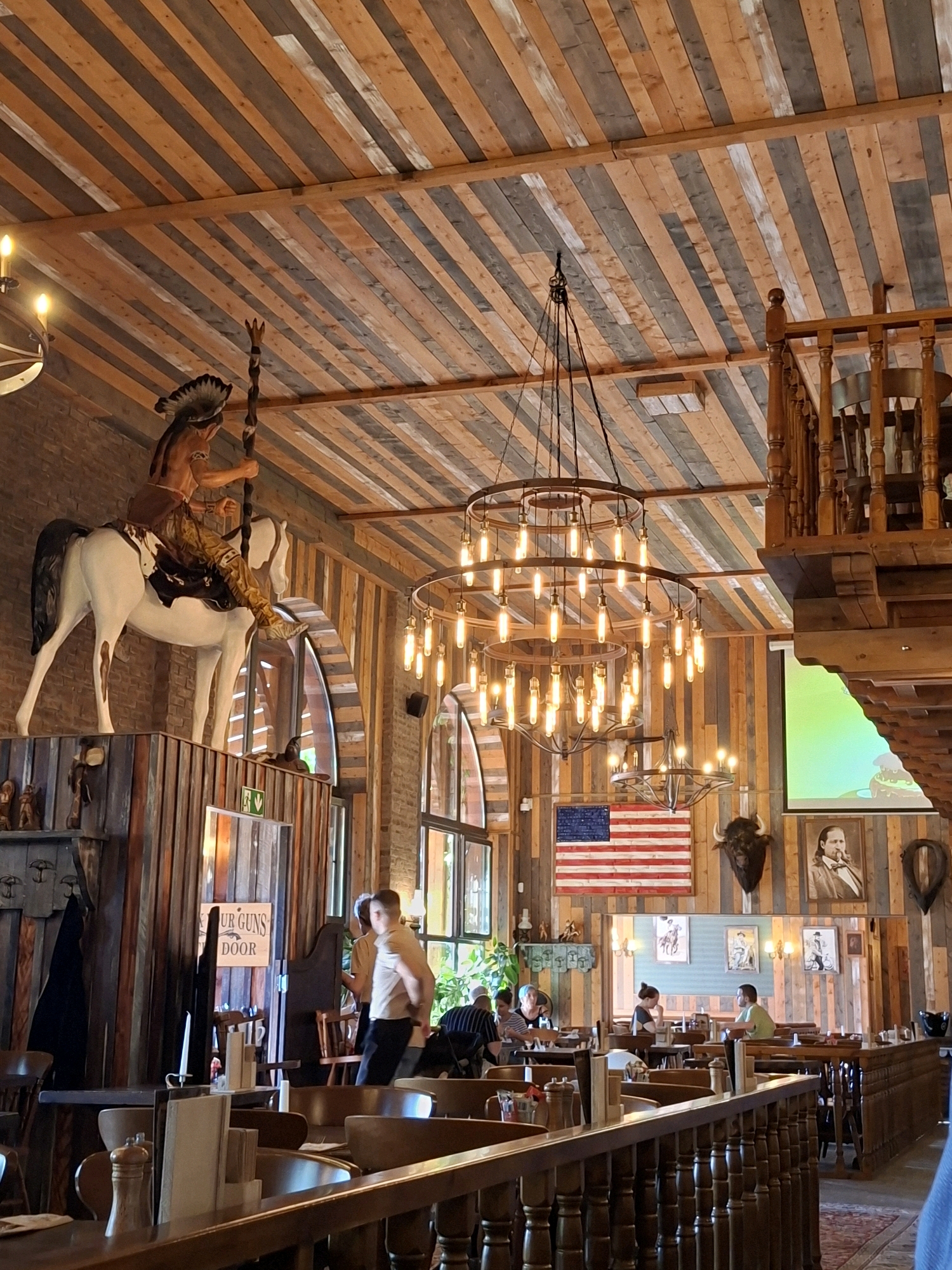 The image depicts the interior of the "Saloon" restaurant in Rastatt, Germany, a themed establishment with a Western American motif. The scene is dominated by rustic wooden elements, including a planked ceiling, wooden walls, and furniture. A prominent feature is a large chandelier with numerous exposed bulbs, casting a warm glow.
In the upper left, a statue of a Native American warrior on a white horse is mounted on a wooden platform, a classic representation of the Wild West. Below this, a sign reads "OUR GUNS DOOR", suggesting a playful theme. An American flag hangs on the wall, flanked by a buffalo head and a portrait of a man in historical attire. Further into the restaurant, multiple tables with chairs are set up, some occupied by patrons. In the foreground, a wooden railing overlooks a dining area. The overall ambiance suggests a lively and immersive dining experience.