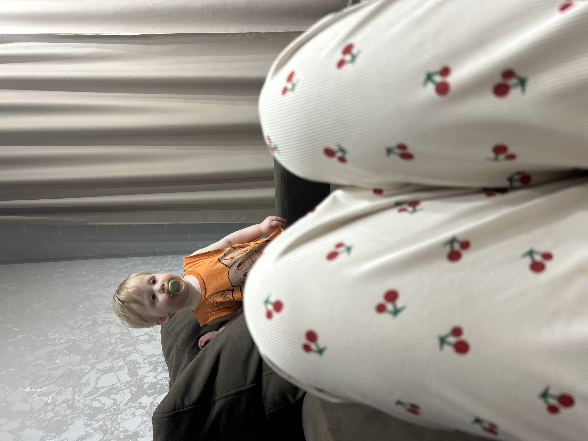 A toddler with blond hair and a pacifier lies on a dark brown couch, looking up and to the left of the frame. The toddler wears an orange shirt. In the foreground, a person's legs clad in white pants with a cherry pattern are positioned above the child. The background features a light-colored wall or curtain. The location is Chaikovsky, Russia.