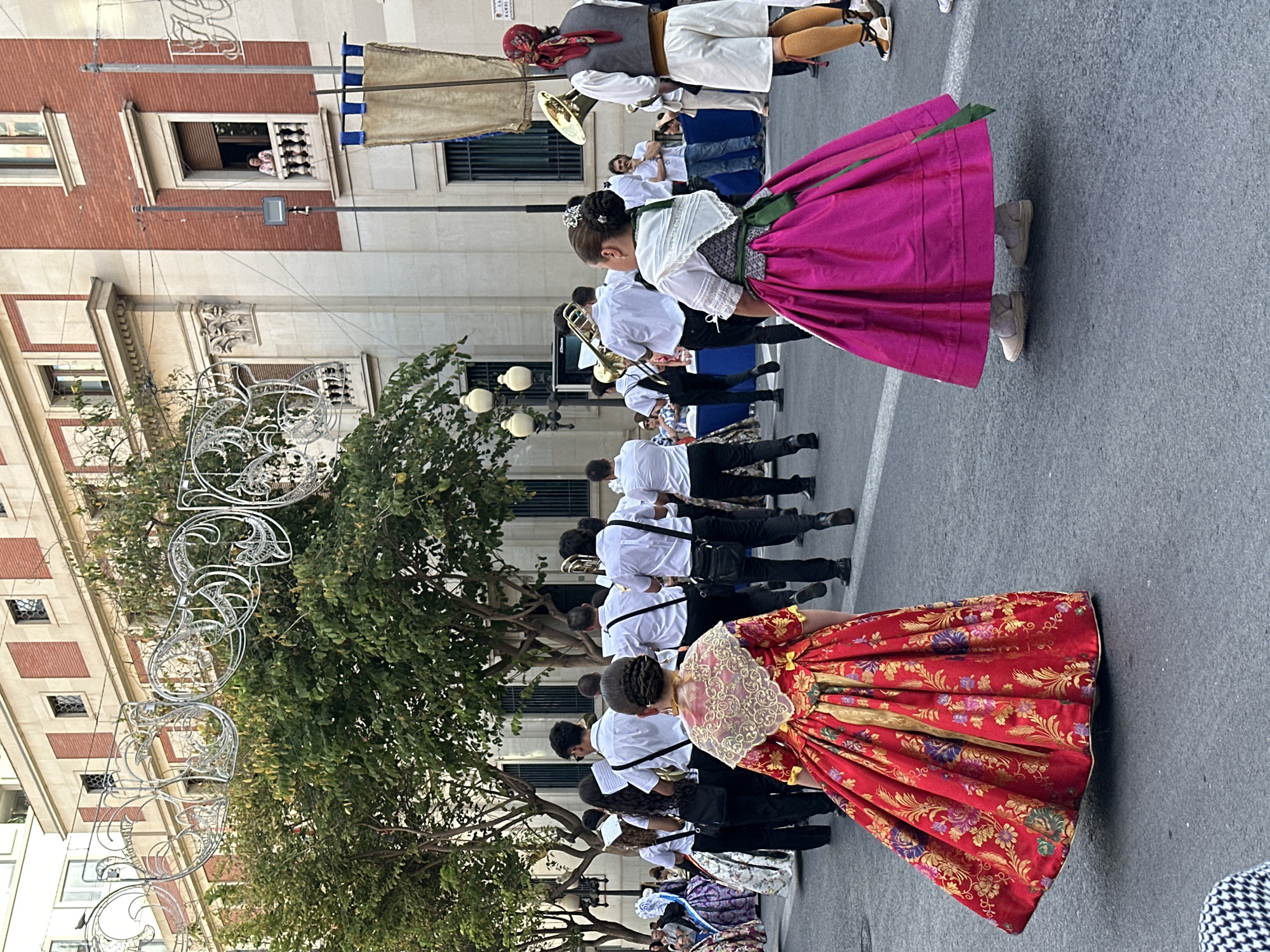 The image captures a street scene in an urban environment, likely a parade or procession. In the foreground, two young women are dressed in elaborate traditional attire. The woman on the left wears a vibrant red gown adorned with a rich floral pattern and gold accents. She has an intricate lace collar and her dark hair is styled in an updo. To her right, another woman wears a bright pink skirt paired with a white embroidered bodice and a green sash. Her hair is also styled up, decorated with a jeweled ornament.
Behind them, a brass band is marching, composed of men and women in white shirts and dark pants. They are holding various brass instruments, including tubas and trombones. Some are looking at sheet music. Further in the background, a building with classical architectural elements, featuring red brick and stone detailing, lines the street. A person can be seen looking out from a second-story window. Decorative street lights and a large banner hang above the street. Spectators are visible in the background on the sidewalk. The overall atmosphere suggests a cultural event or festival taking place in a city square or street.