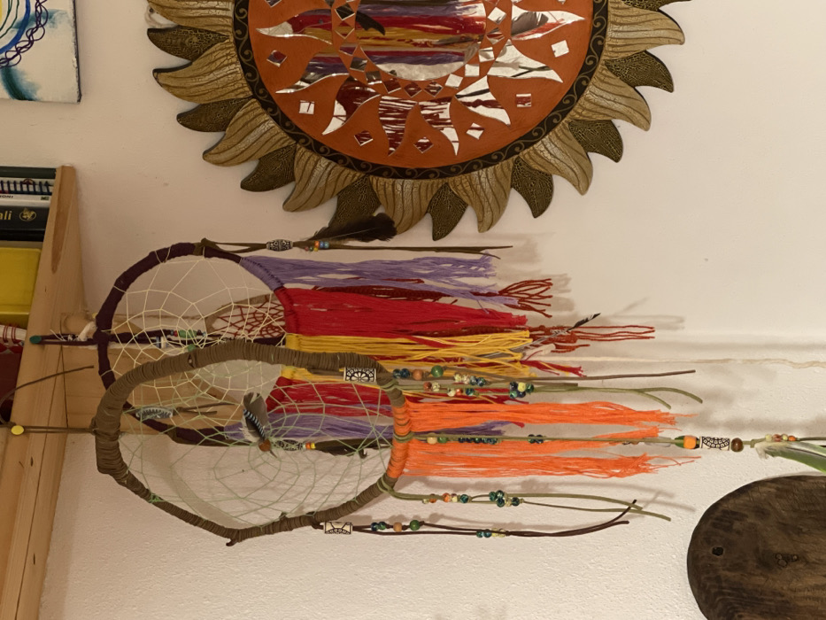 The image captures a collection of decorative objects mounted on a light-colored wall, likely in an interior space within Torre di Santa Maria, Italy.

At the top, a large, intricate sun-shaped wall hanging is prominently displayed. It features an orange central disc embedded with small mirror pieces, surrounded by textured, golden-brown rays.

Below the sun decor, several dreamcatchers are visible. They vary in size and design; one has a dark purple-wrapped hoop, and another a light brown-wrapped hoop, both containing delicate white web-like netting. Hanging from these dreamcatchers are numerous colorful tassels made from yarn in shades of purple, red, yellow, and orange, interspersed with green and brown strings. These hanging elements are adorned with a variety of beads in green, orange, brown, white, and multicolored patterns, along with small silver-colored metal accents. Brown and spotted feathers are also integrated into the dreamcatcher designs.

To the far left, a section of a light-colored wooden shelf or furniture piece is visible, supporting what appear to be book or box spines with partially readable text, including "ali" and "manu." A small corner of a white canvas artwork with blue circular patterns is visible in the top left. In the bottom right, a portion of a dark, round wooden object is present against the wall. No human activity is depicted; the scene portrays static decorative items, creating a warm, bohemian, and artfully arranged atmosphere.