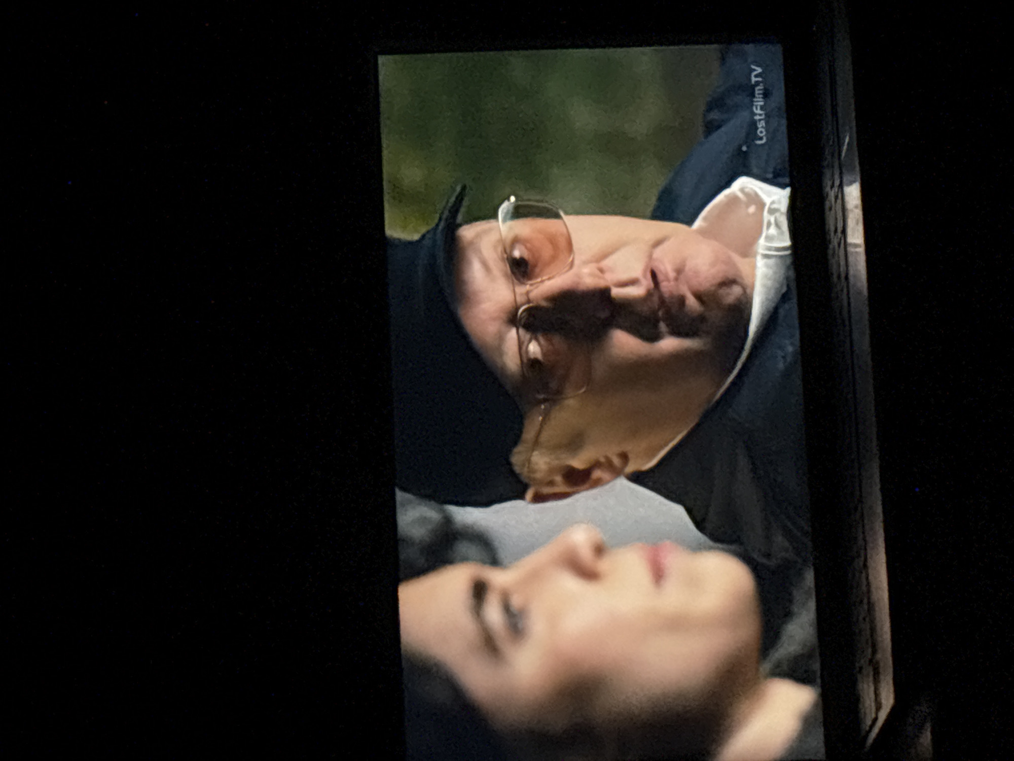 The image depicts a scene from a video, displayed on a screen. The screen is partially framed by a dark object, likely part of a phone or camera. In the video, two people are visible. The person in the foreground is a woman, lying down with her face upturned. Her features are softly blurred, but she appears to be young. Above her, another person, likely a man, is leaning over her. He is wearing glasses and a dark cap, with a light-colored shirt visible beneath a darker jacket. The background is blurred, suggesting an outdoor setting, potentially at night. The logo "LostFilm.TV" is visible in the top right corner. There is no information about Budva, Montenegro.